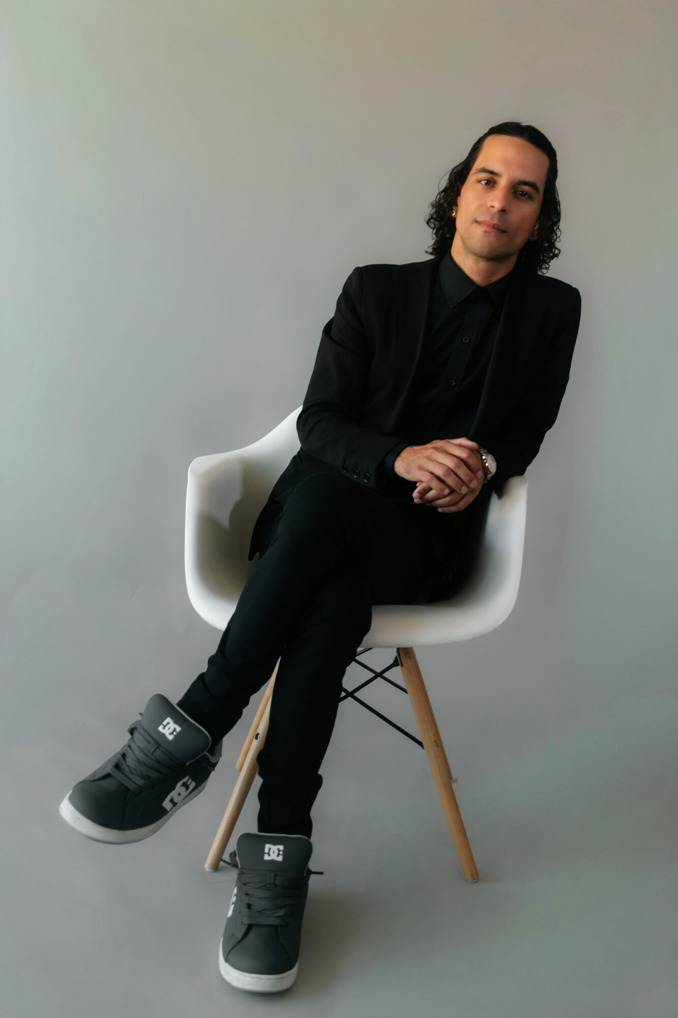 Person with long dark hair sitting on a white chair against a plain background, wearing dark clothing and sneakers.