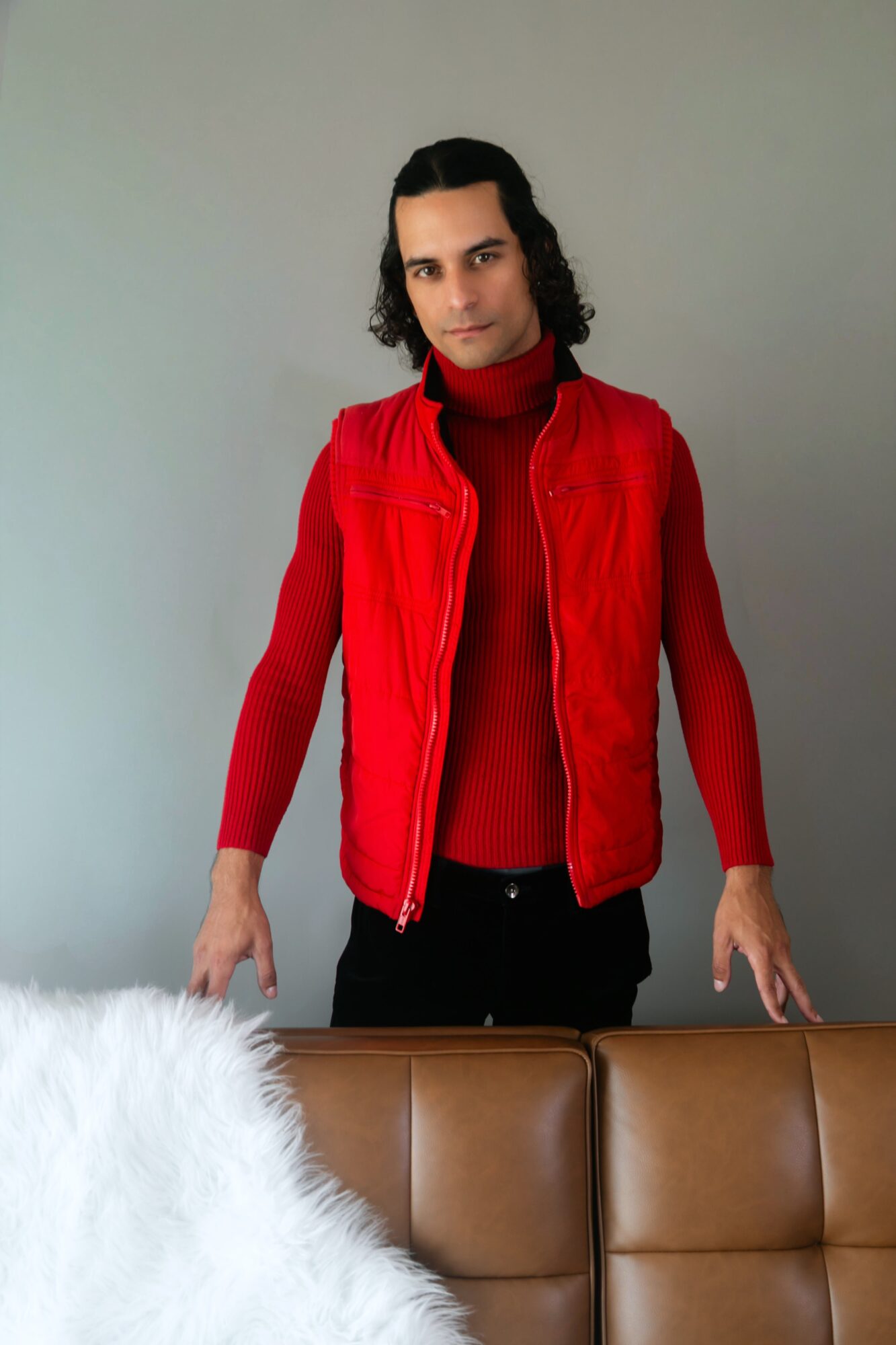 Person with shoulder-length dark hair wearing a red vest and red turtleneck sweater, standing behind a brown sofa.