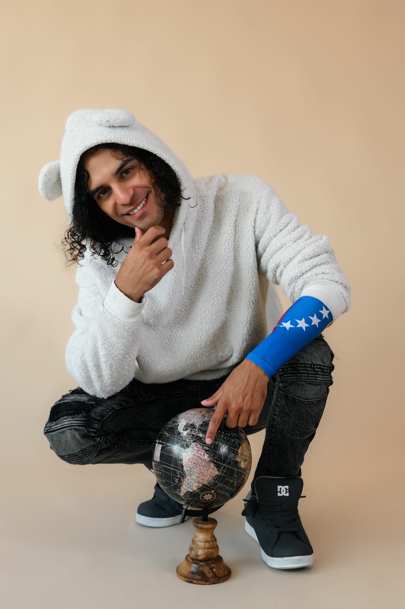 Person crouching with a disco ball, wearing a white fuzzy hat and sweater, smiling, against a plain background.