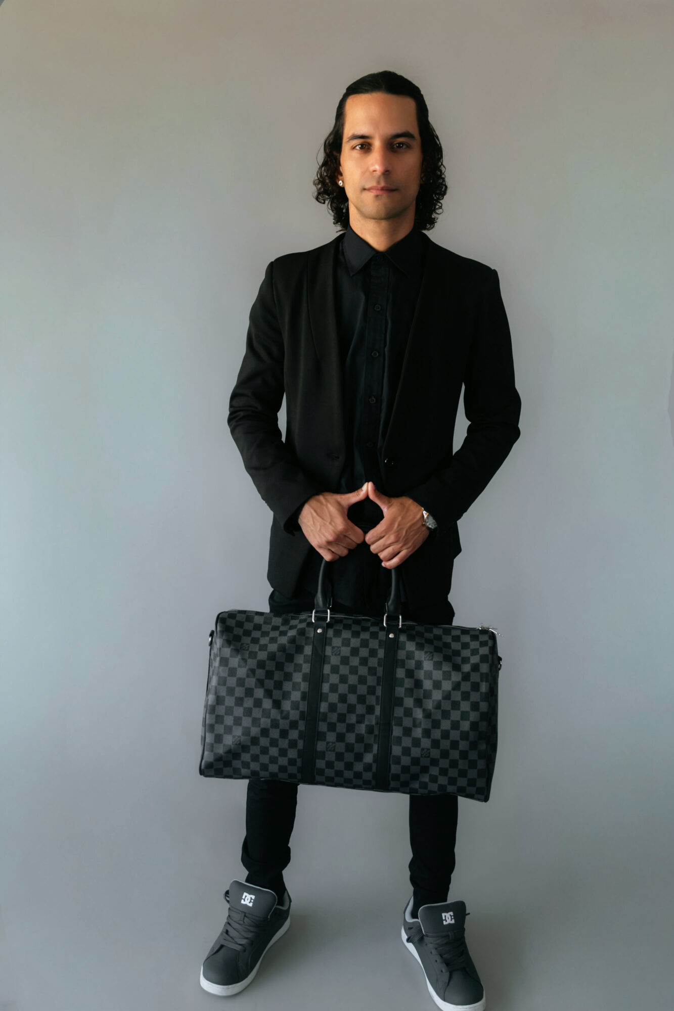 Man in black suit holding a checkered bag, standing against a plain light background.