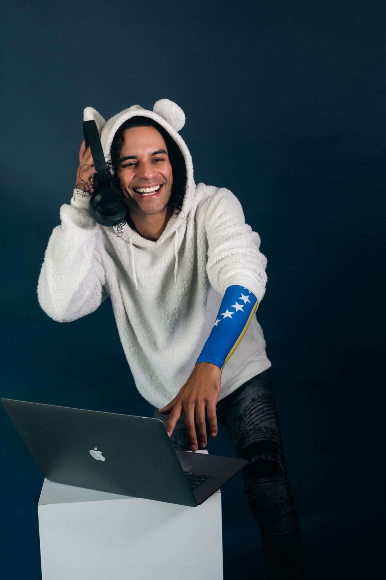 Person wearing a hoodie and headphones, smiling, with a laptop on a white stand against a dark background.