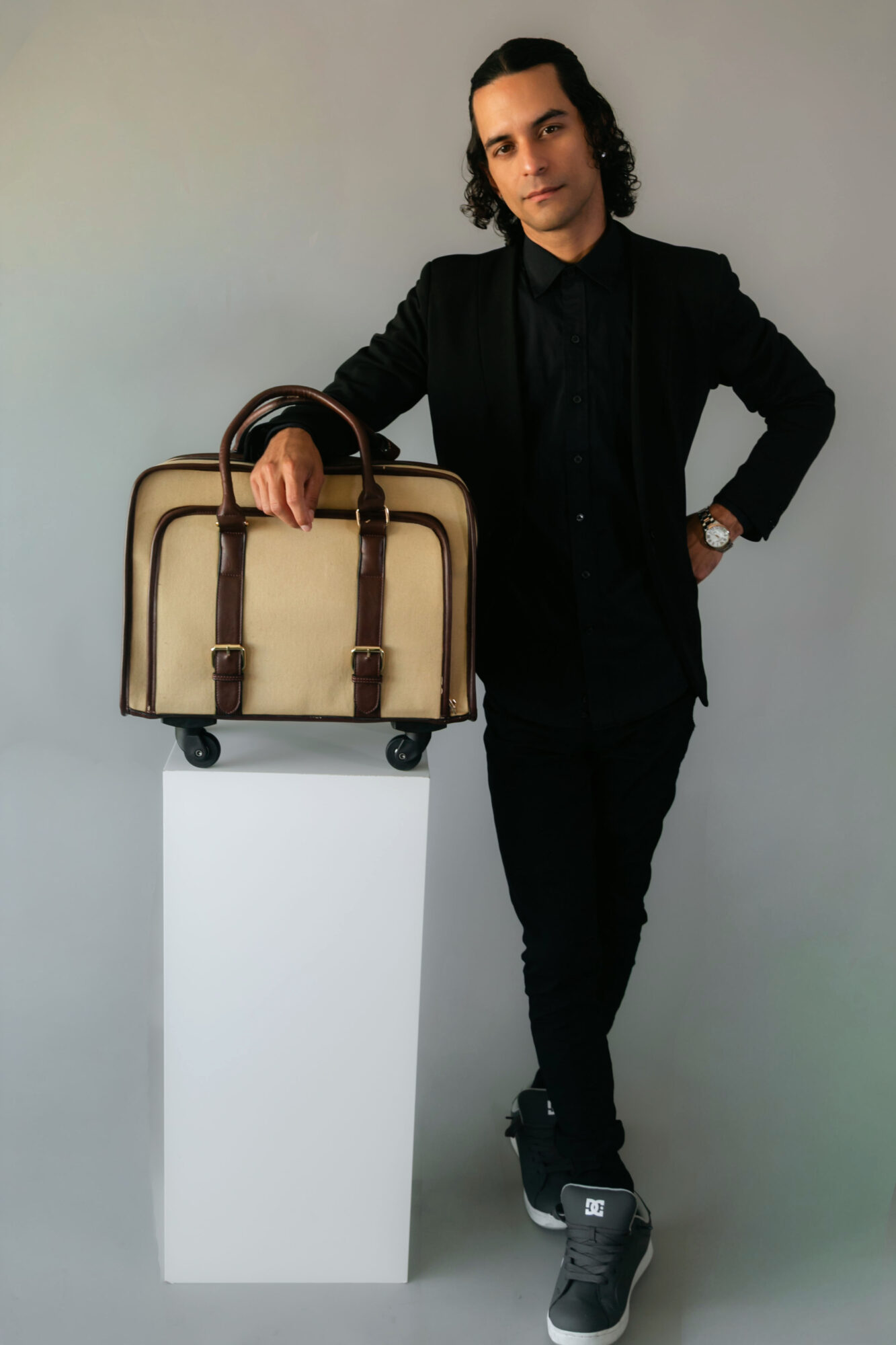 Man with long dark hair in black suit standing next to a beige and brown suitcase on a white pedestal, against a plain background.