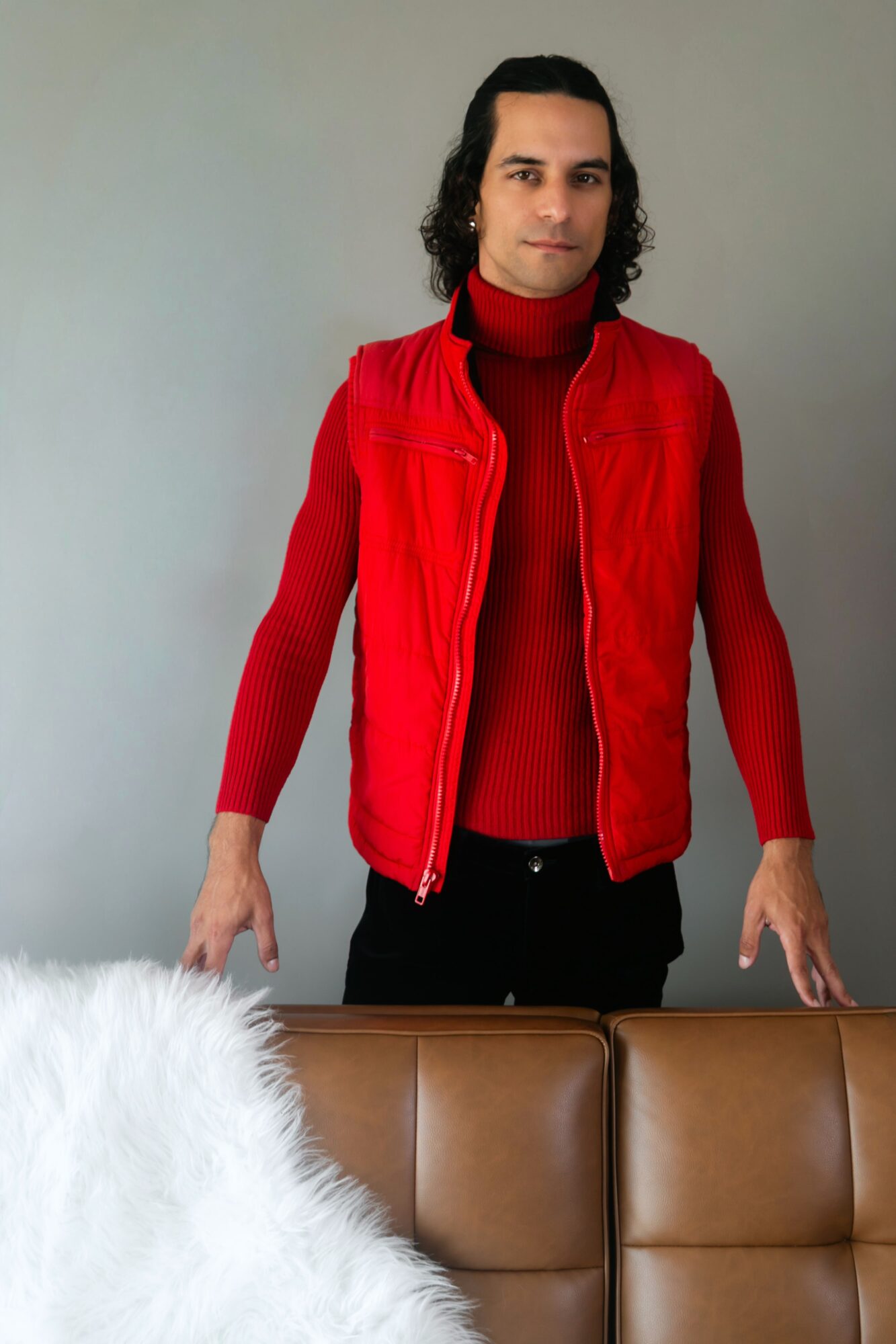 Person with long dark hair wearing a red jacket and turtleneck sweater, standing behind a brown sofa, against a plain wall.