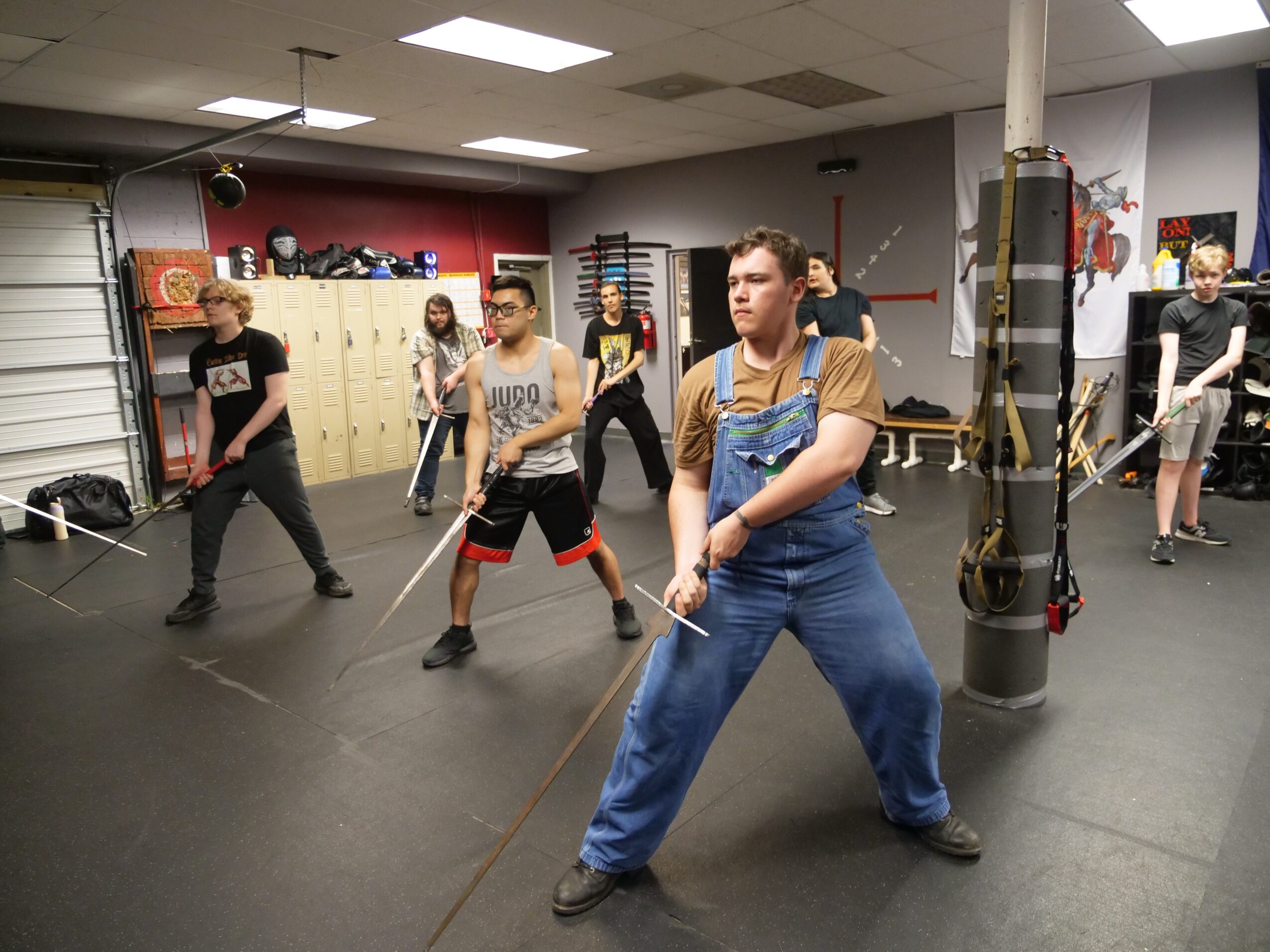 Group of people practicing fencing in a gym with lockers and equipment, some wearing protective gear.
