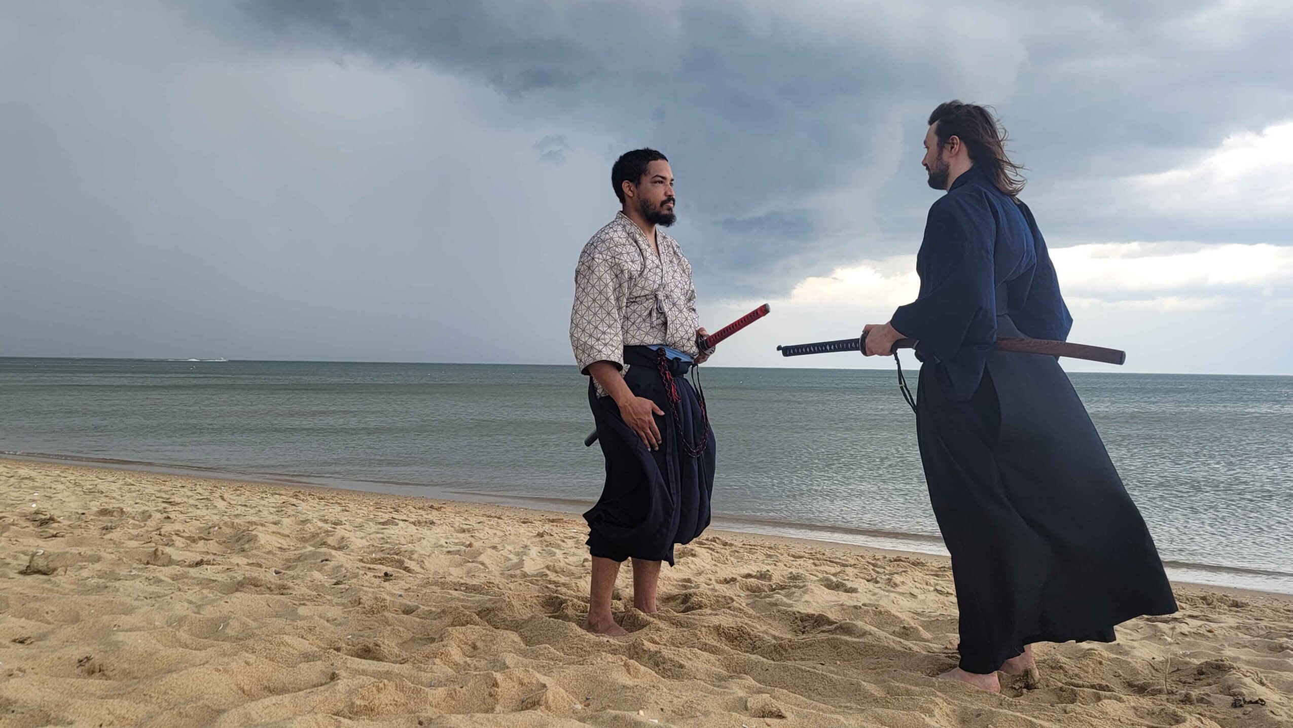 Two people in traditional clothing stand on a beach, facing each other with a cloudy sky and ocean in the background.