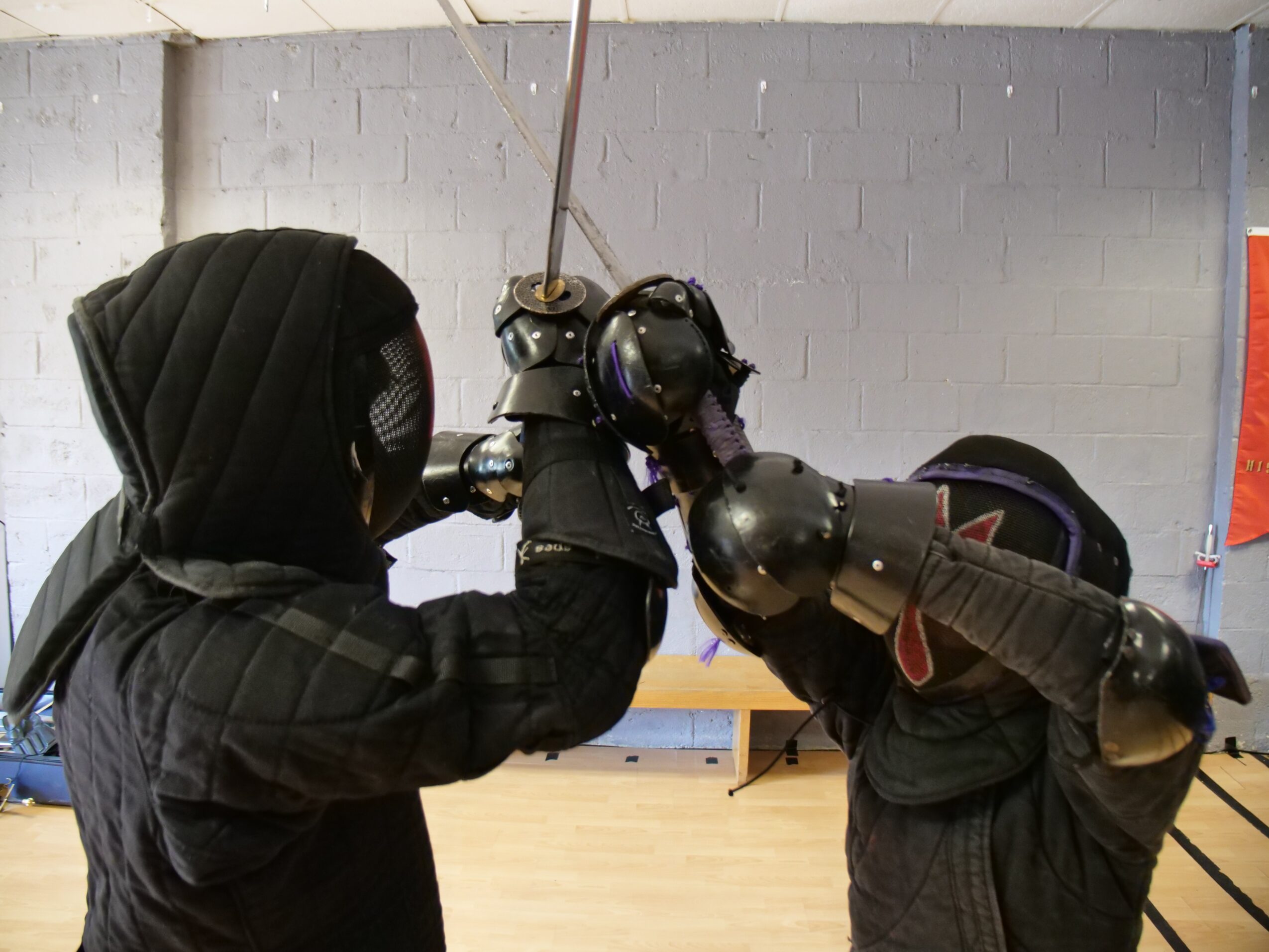 Two individuals wearing black hooded clothing and helmets, one with a shield, engaged in a confrontation indoors.