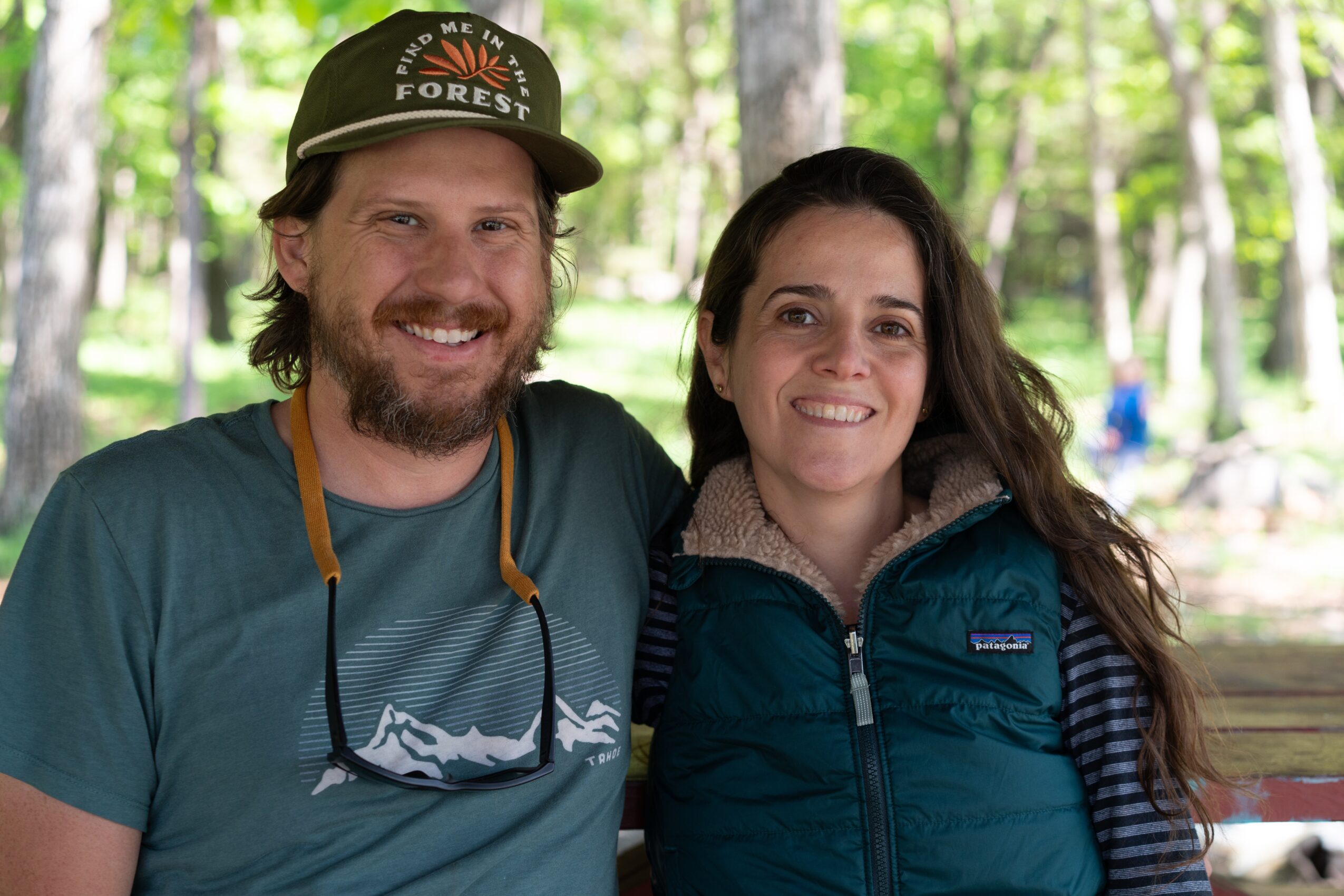 Two people smiling outdoors in a forest, one wearing a cap and the other in a jacket.