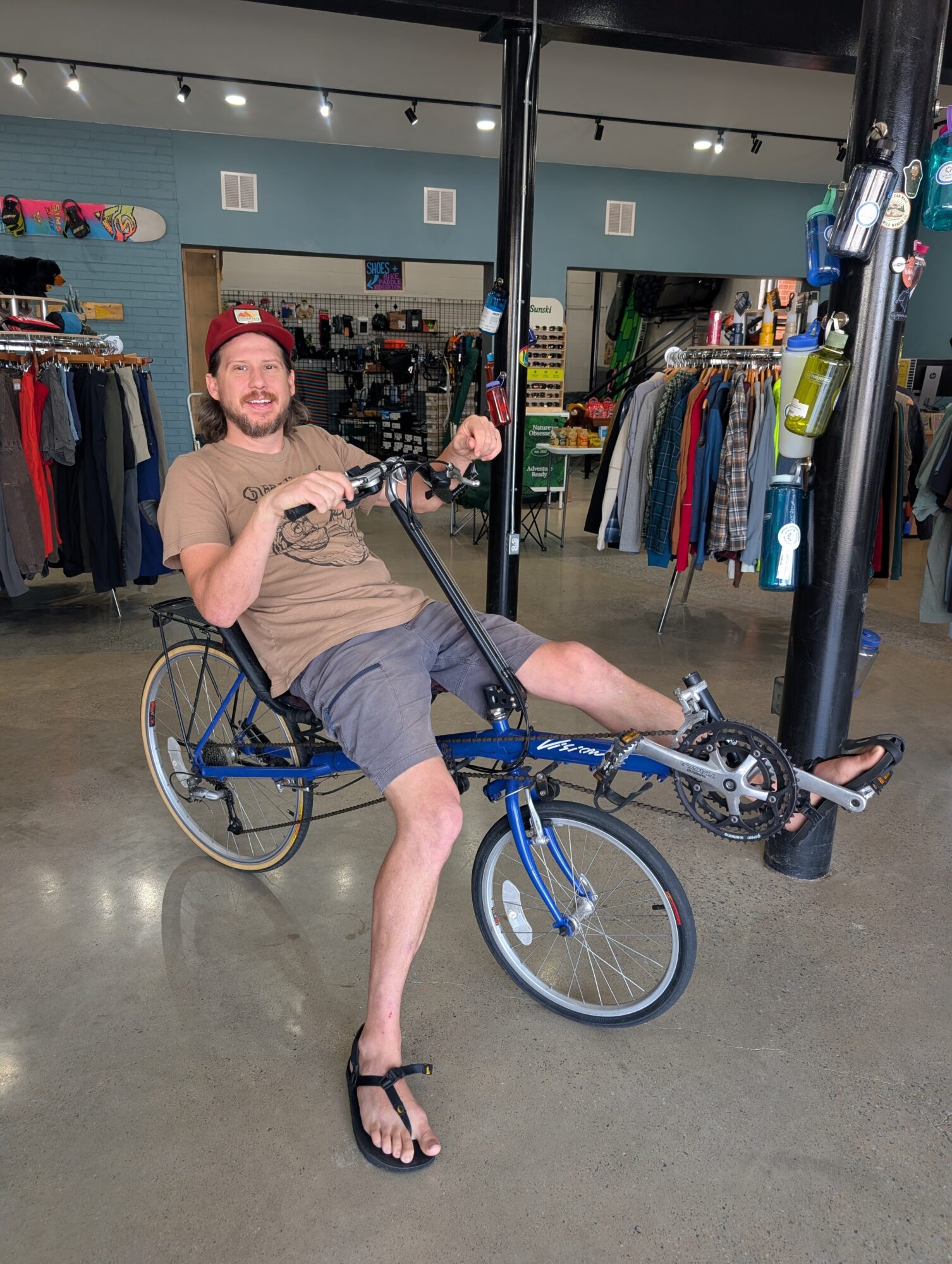 Person in a wheelchair with a bicycle frame, smiling indoors with clothing racks in background.