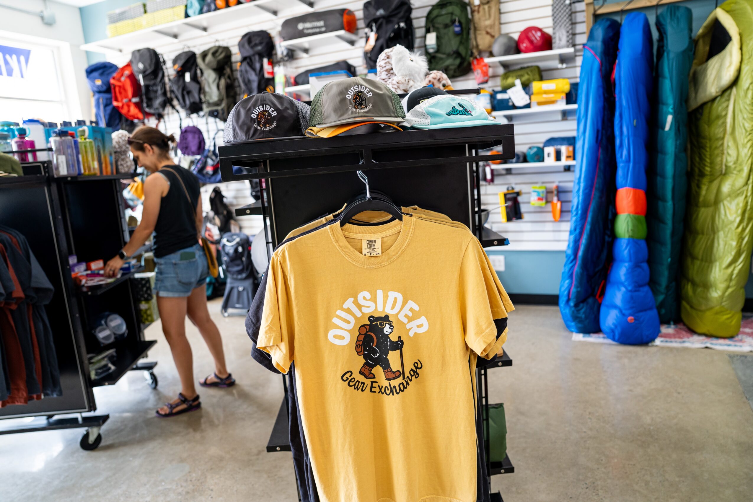 Yellow T-shirt with a bear graphic and text, hanging in a store with outdoor gear and clothing in the background.