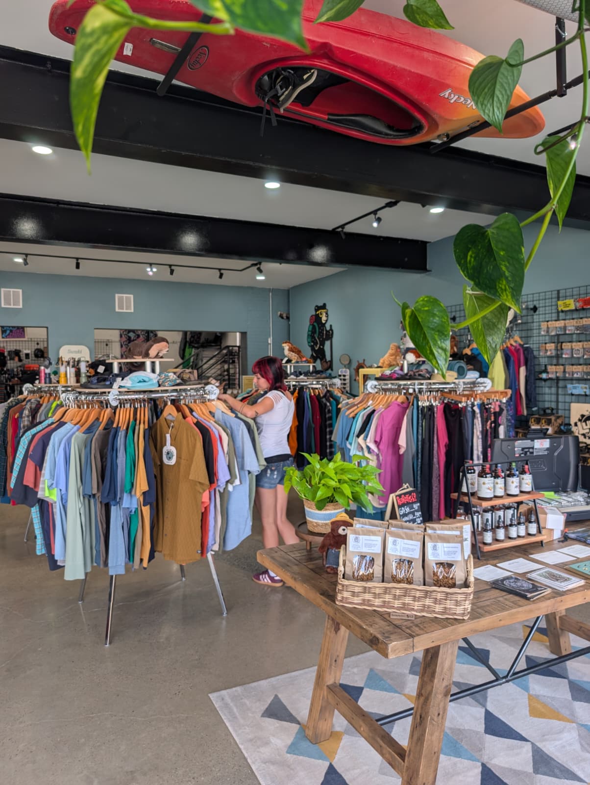 Clothing store with racks of colorful shirts, a customer browsing, and shelves with accessories, plants, and display tables.