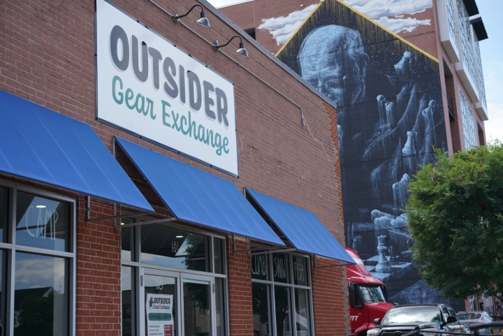 Storefront with blue awnings and a sign reading 'OUTSIDER Gear Exchange' on a brick building.