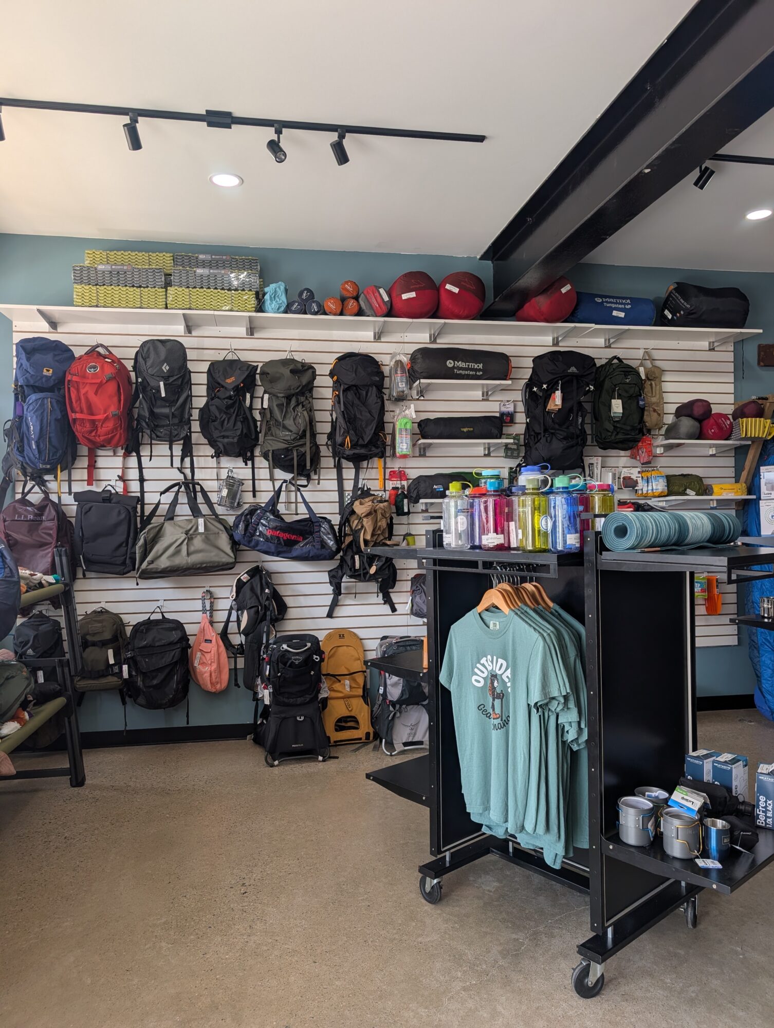 Shelves with backpacks, helmets, and outdoor gear, along with a clothing rack and shelves of supplies in a store.