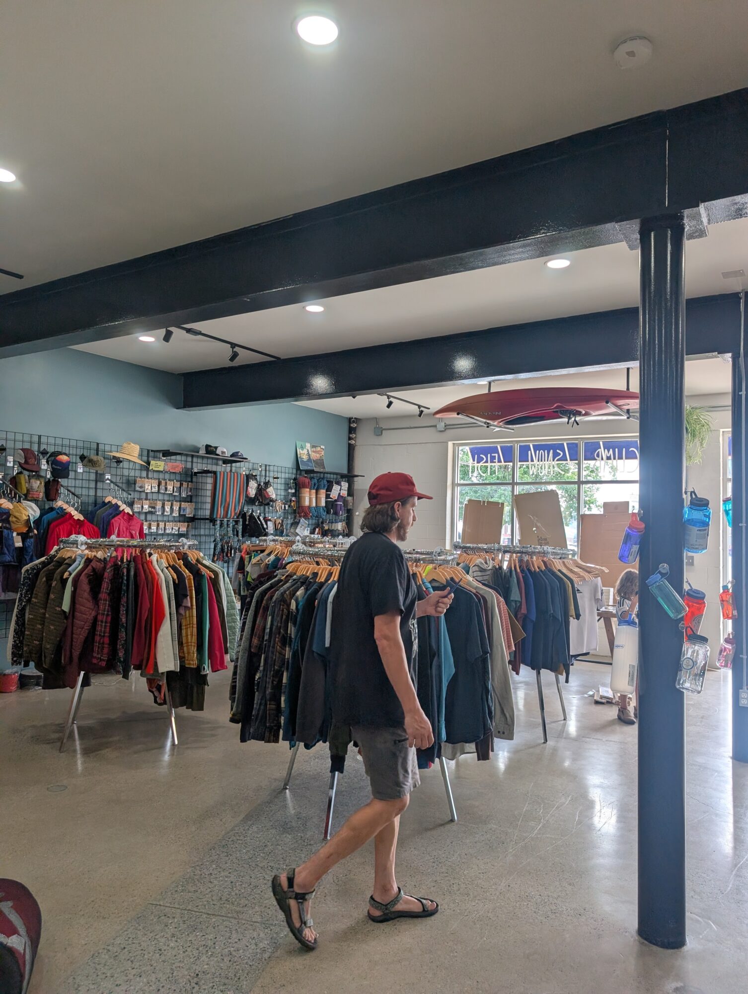 Person walking inside clothing store with racks of clothes, large windows, and a red umbrella outside.