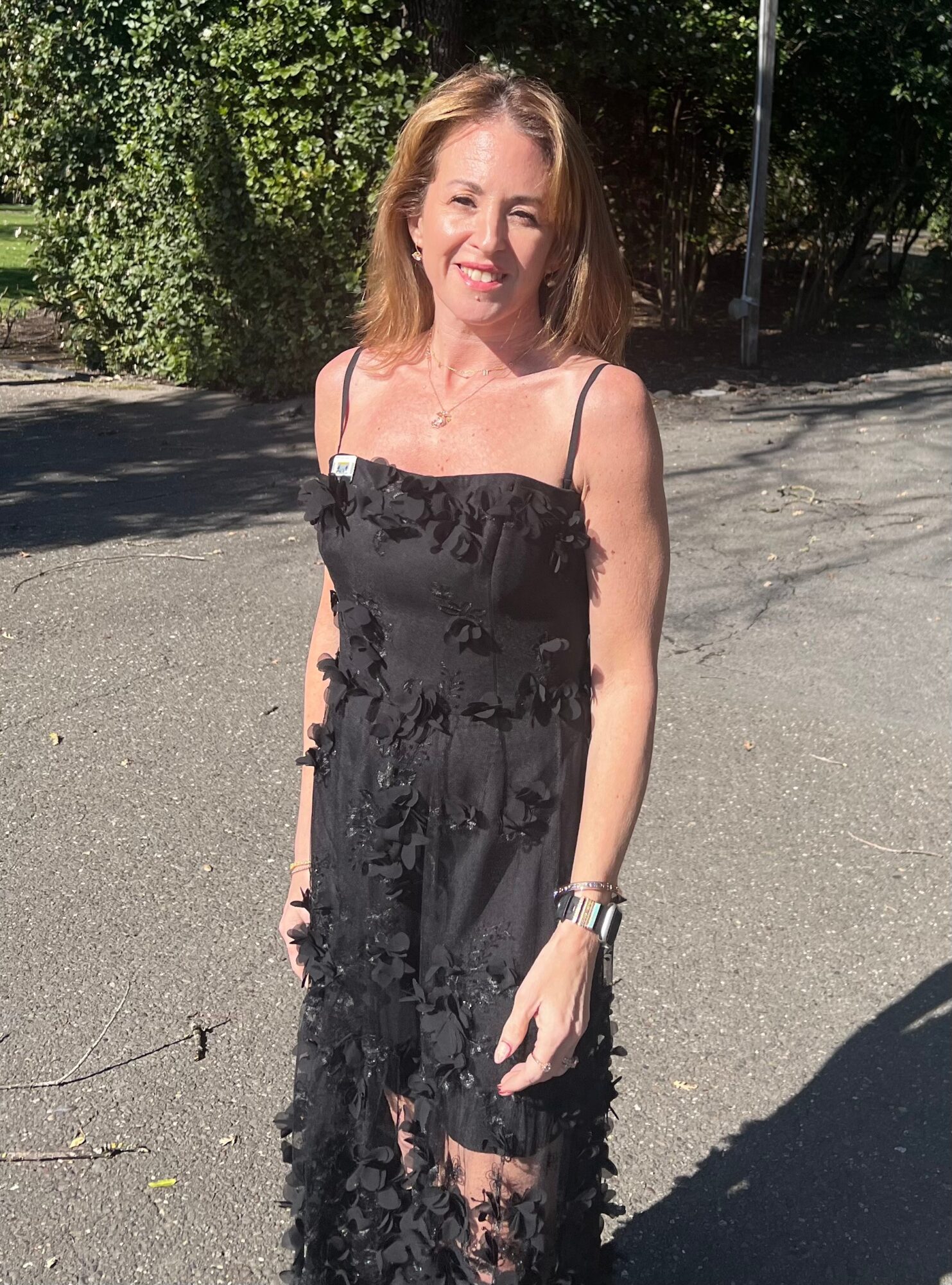 Woman standing outdoors on a sunny day, smiling, wearing a black dress with spaghetti straps, with trees in the background.
