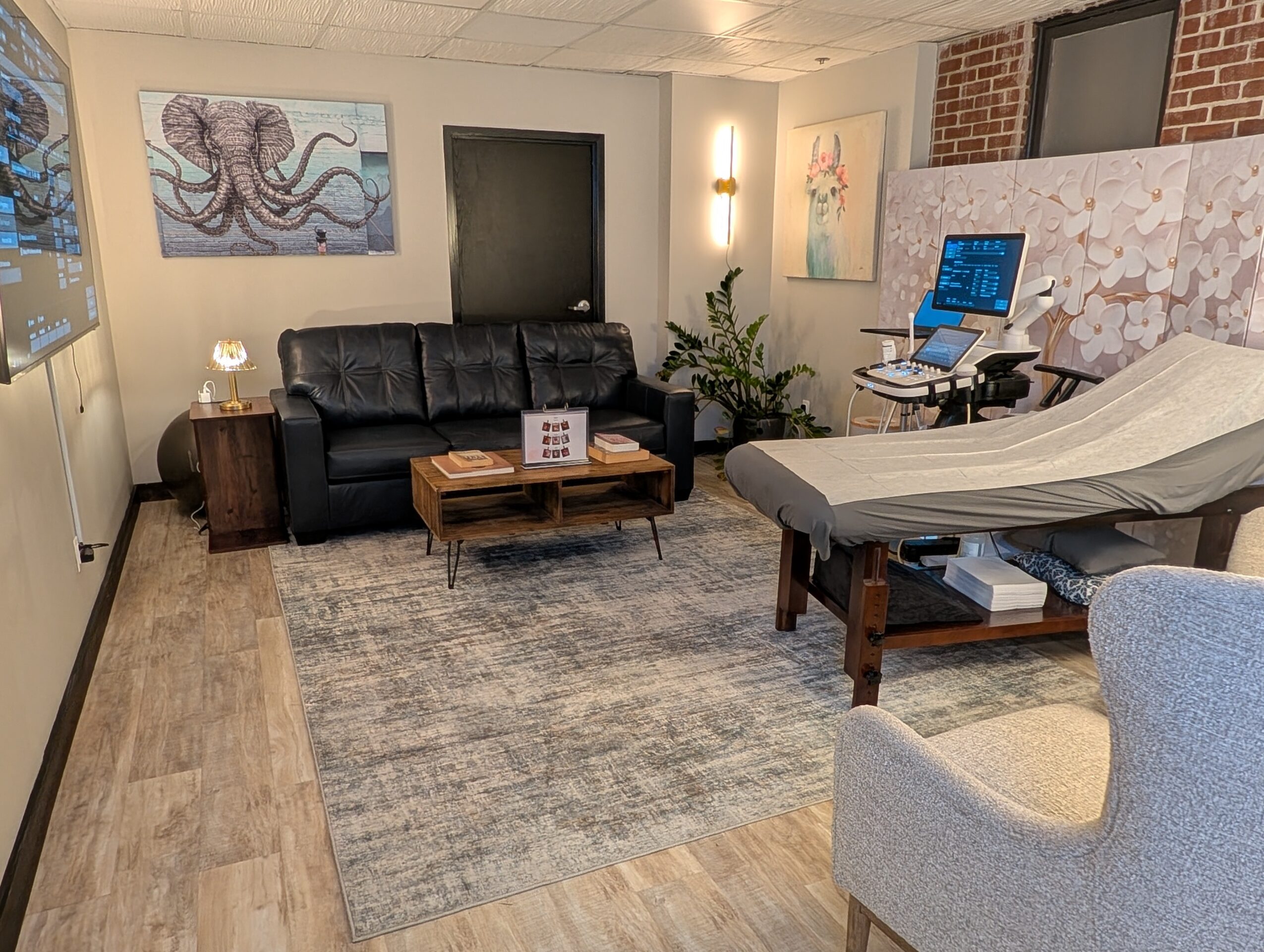 Medical examination room with a bed, a gray armchair, a black sofa, artwork on the walls, and a computer on a stand.