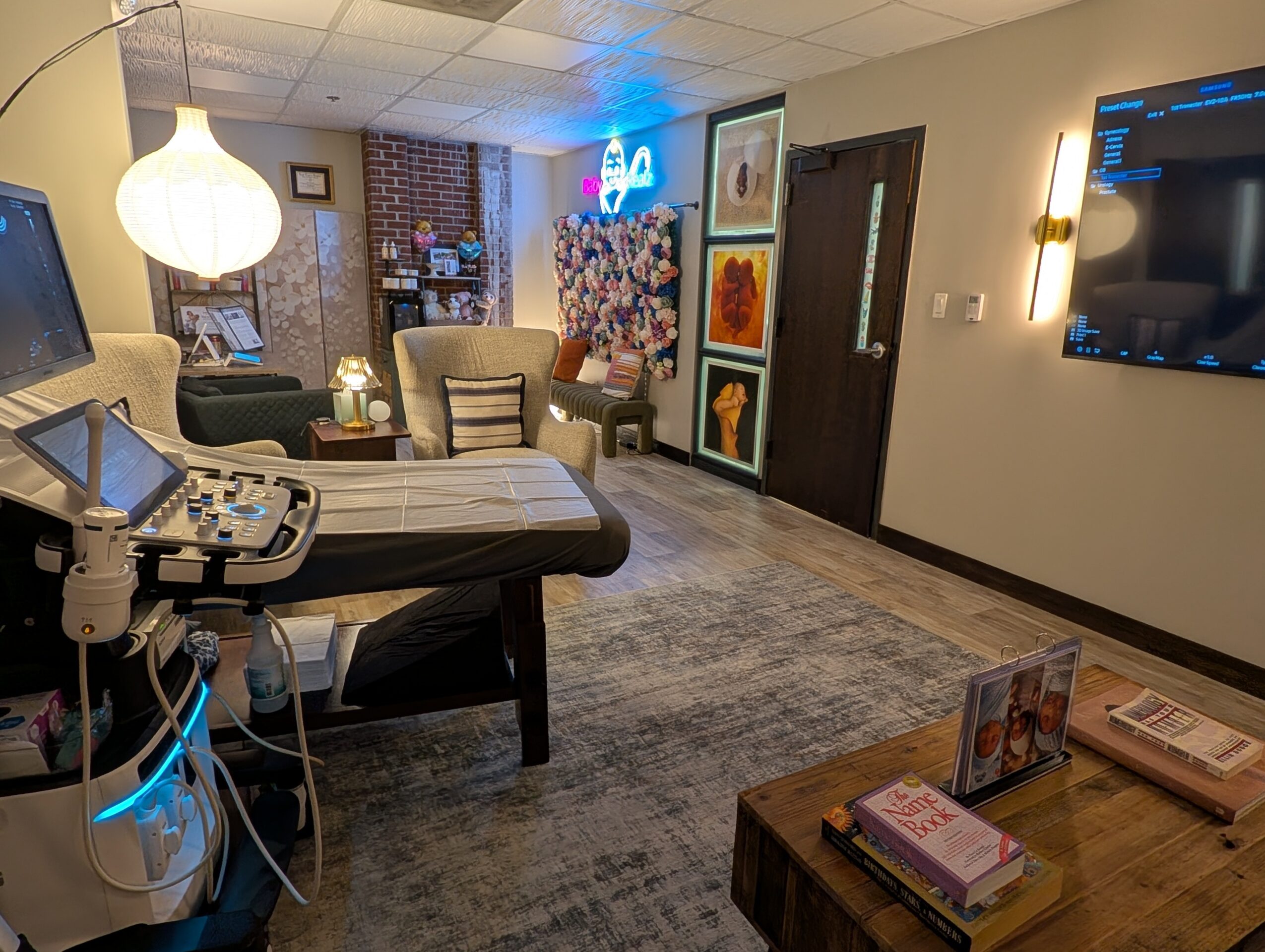 Living room with chairs, a table, wall art, a TV, and a medical ultrasound machine, illuminated by a hanging lamp.