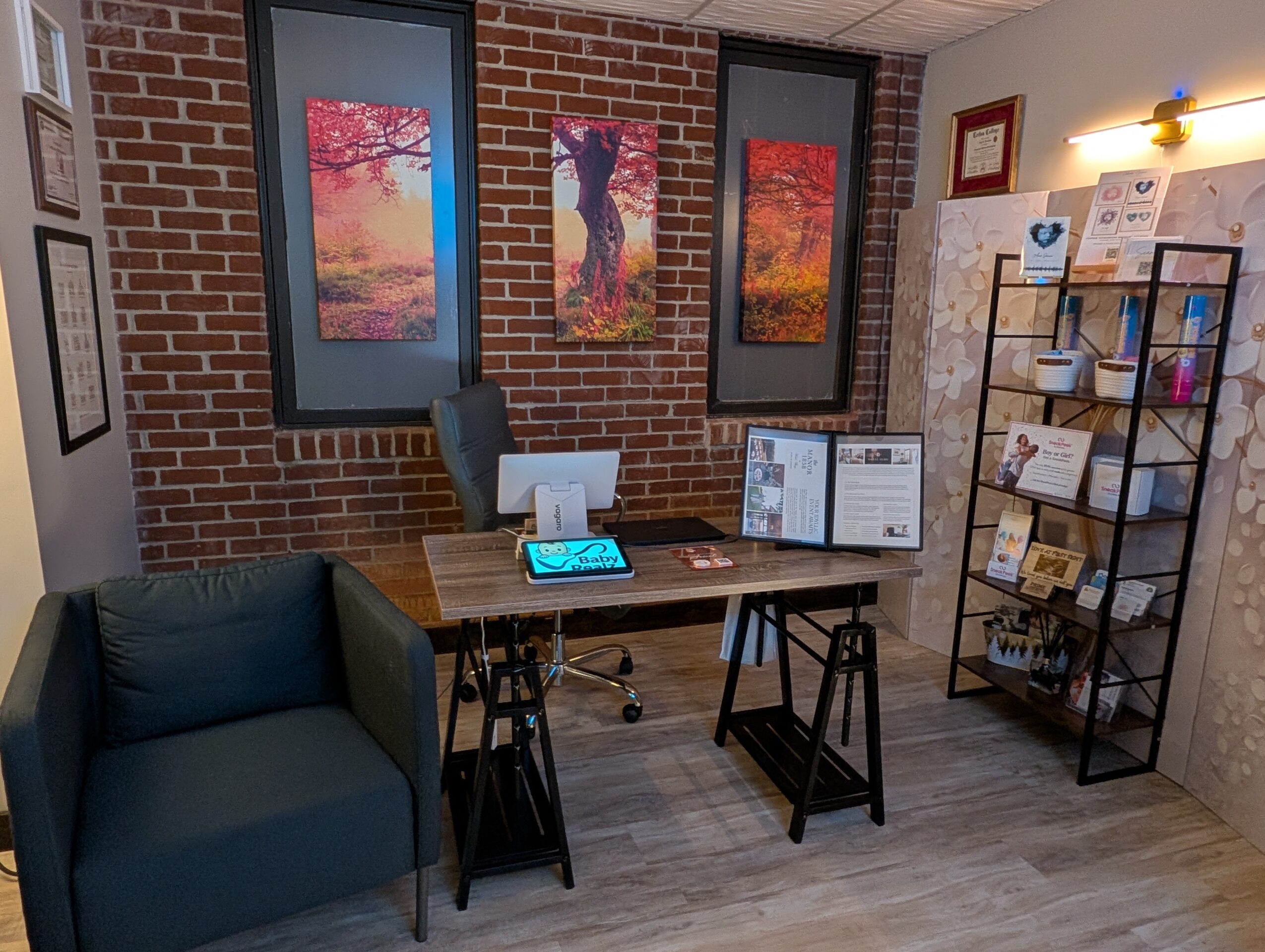 Office corner with brick wall, three colorful landscape paintings, desk with computer, and a bookshelf with brochures.