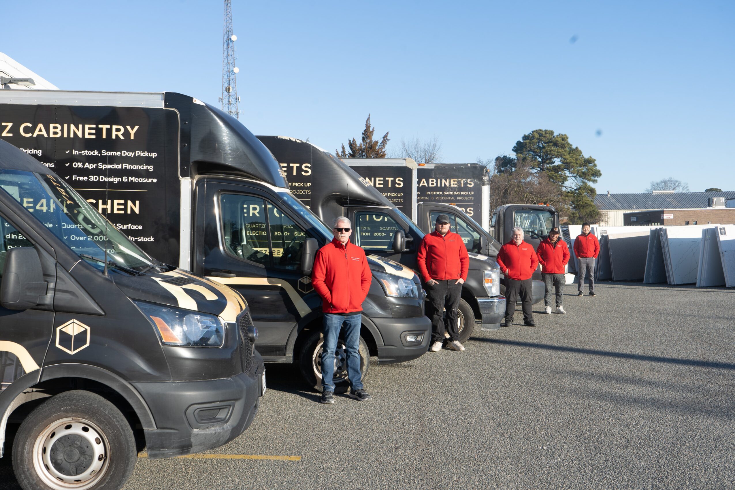 Line of black service vans with company logos and people in red shirts standing outside on a paved lot under a clear blue sky.