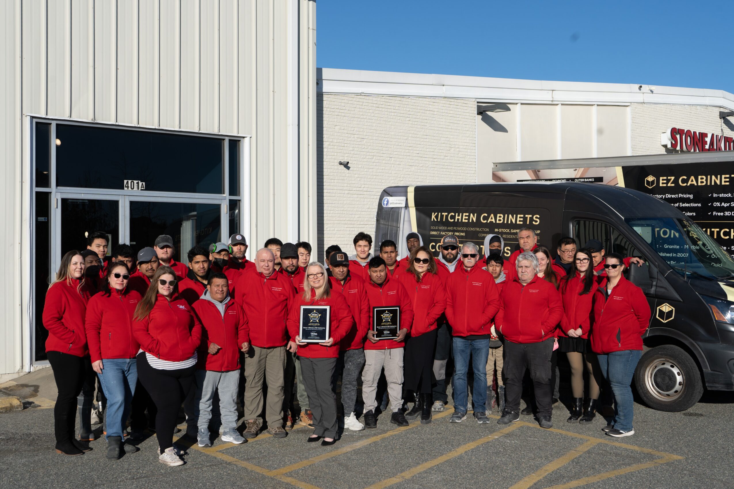 Group of people wearing red jackets standing outside a building with a black van behind them, sunny day.