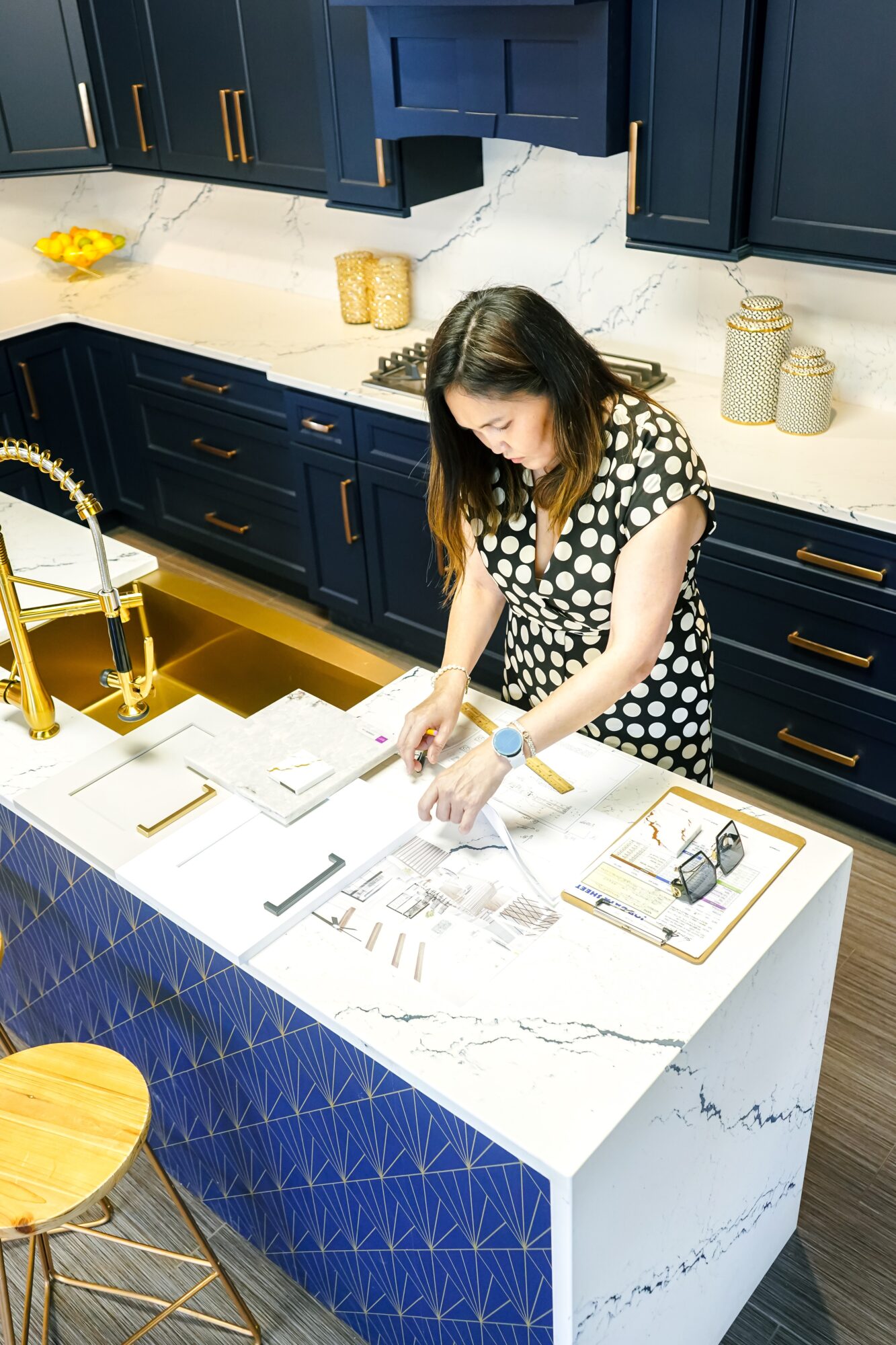 Woman in polka dot dress preparing food on kitchen island with blue cabinets and gold accents, marble countertop.