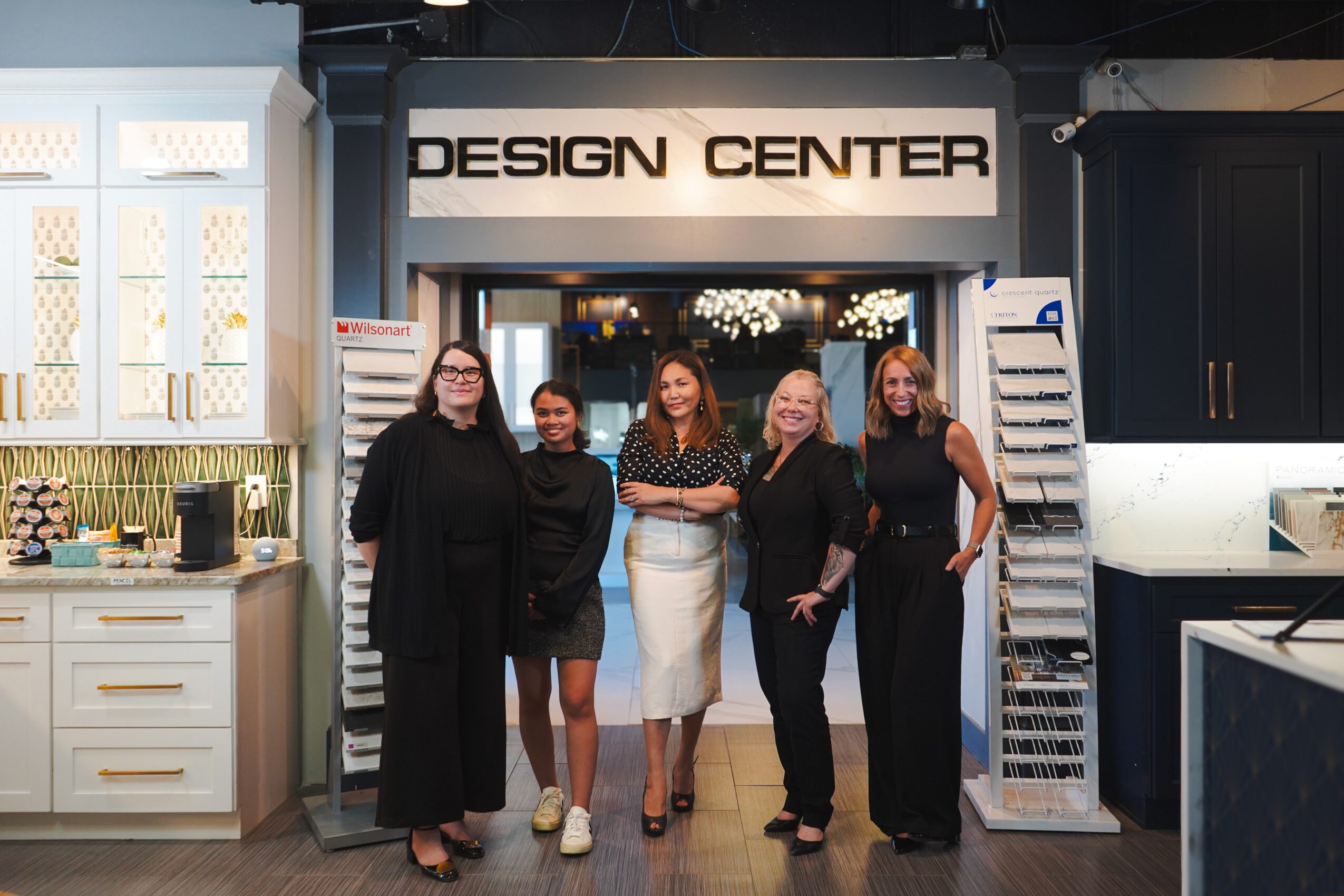 Five women stand in front of a design center entrance, smiling, with display racks on either side, in an indoor setting.