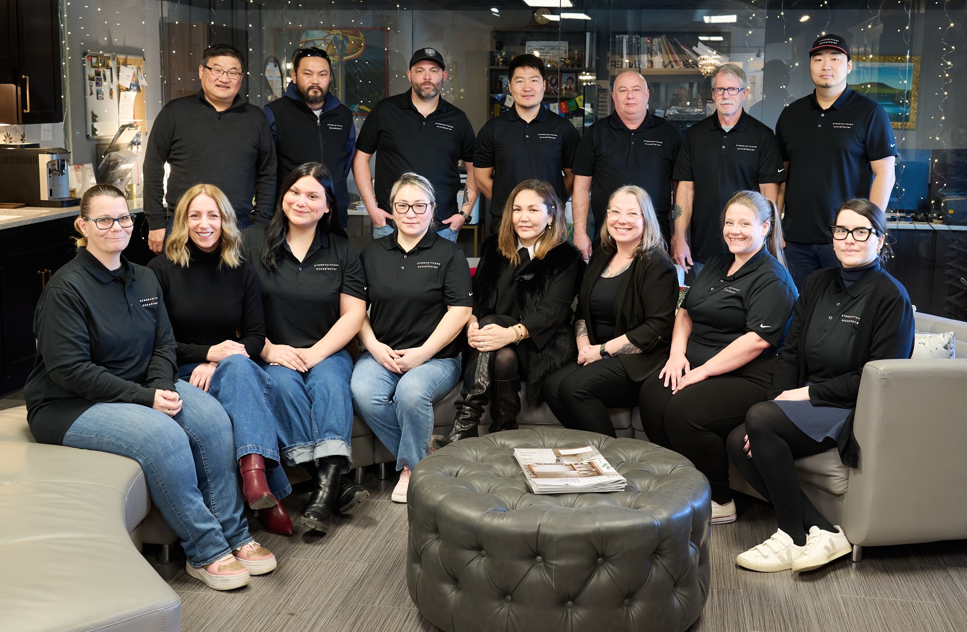 Group of 14 people in black shirts, seated and standing in a modern office, smiling, with a round ottoman and magazines in foreground.