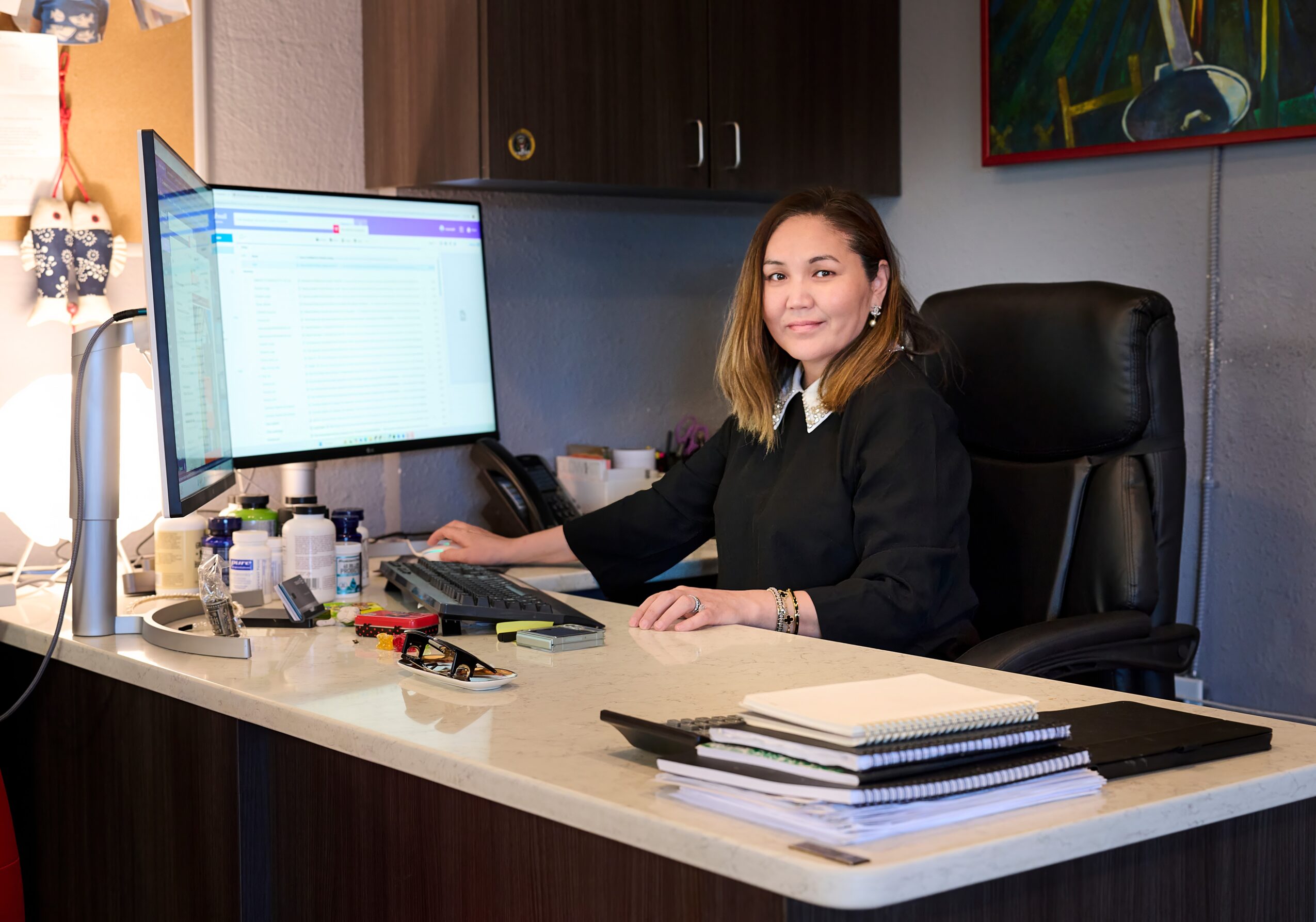 Woman sitting at desk with two monitors, smiling, surrounded by office supplies and documents.