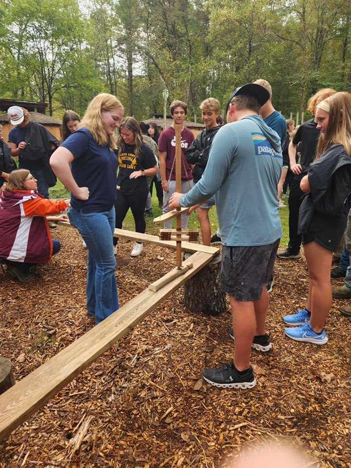 Group of people outdoors around a wooden structure, trees in background, some wearing masks, engaged in activity.
