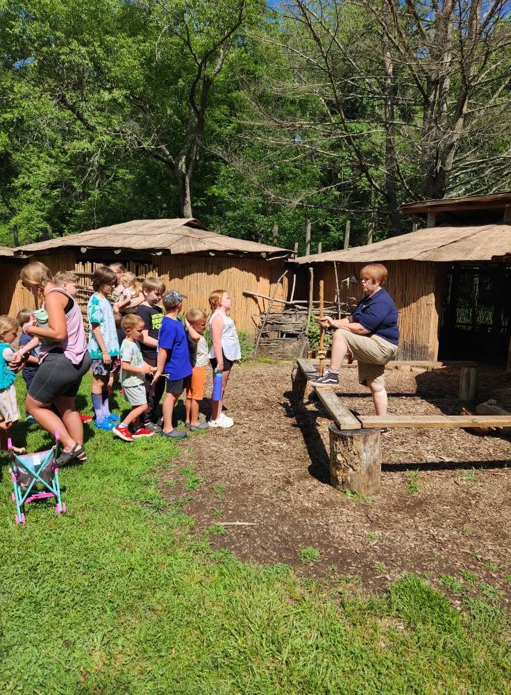 Children gather around a person on a wooden platform outdoors, with trees and rustic buildings in the background.