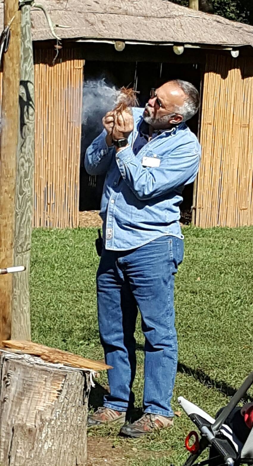 Man with glasses and beard, wearing a blue jacket and jeans, standing outdoors near a wooden structure, holding a cigarette.