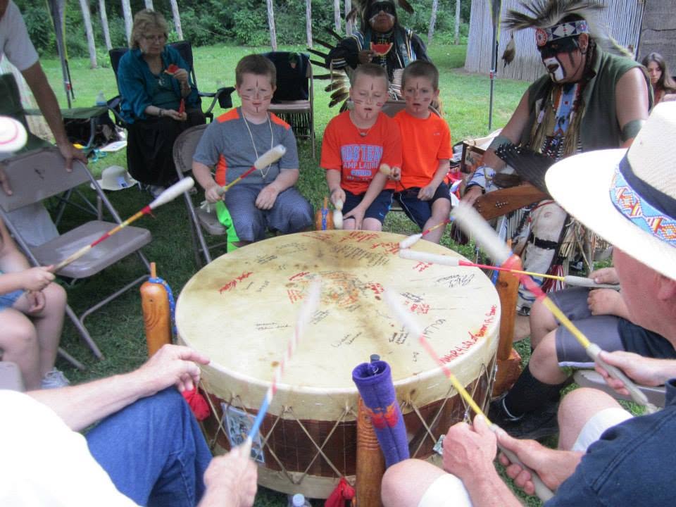 Group of children and adults sitting outdoors around a large drum, playing with sticks, in a grassy area with trees.