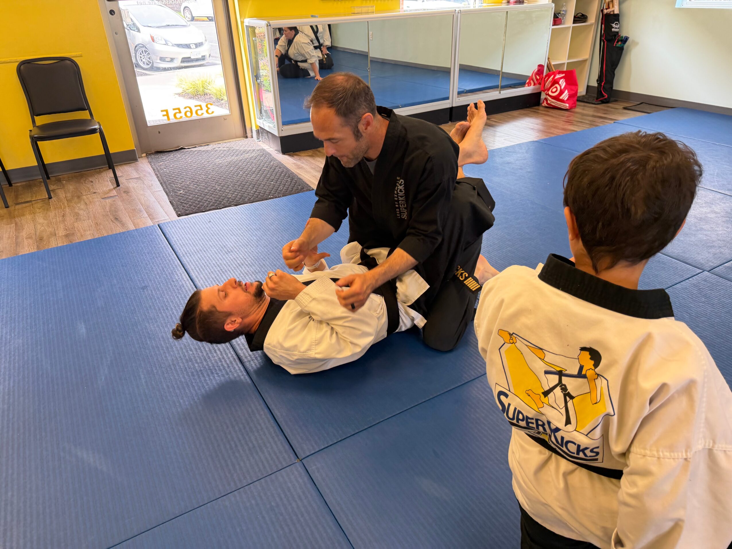 Martial arts instructor practicing grappling techniques with a student on blue mats, with another student observing nearby.