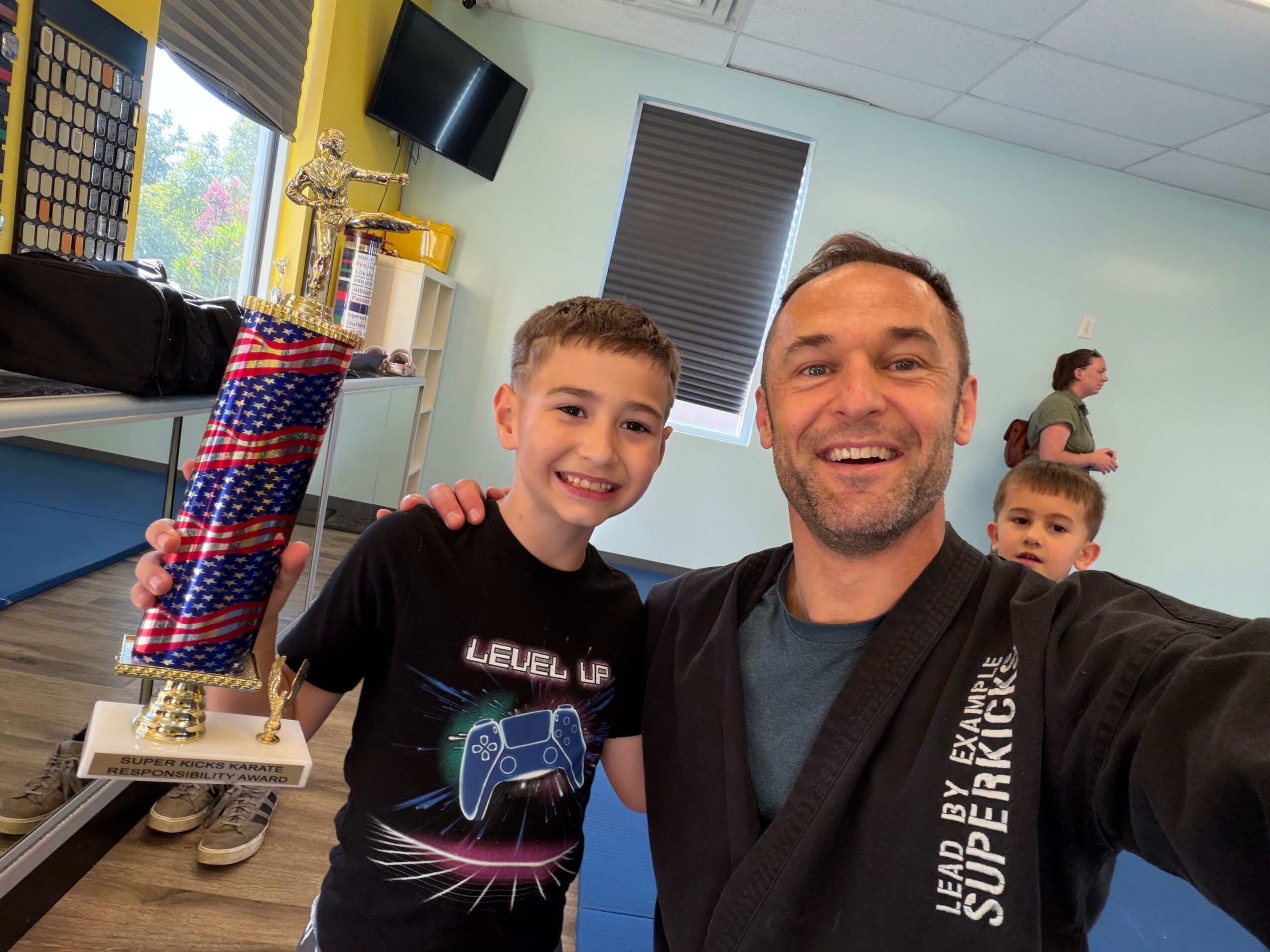 Two people smiling in front of a large colorful cake with candles, in a room with a window and a TV. Word on shirt: 'LEAD BY EXAMPLE SUPERKID'.