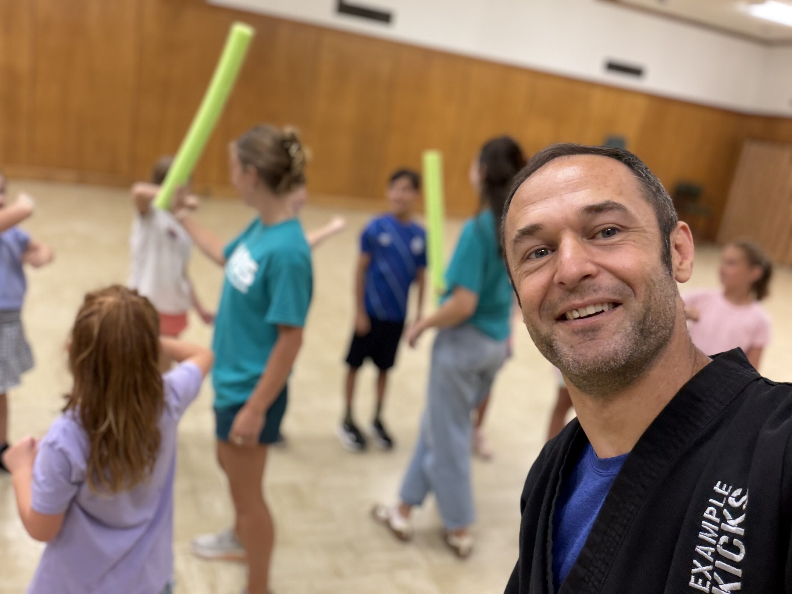 Man taking a selfie with children playing with foam noodles in a gymnasium.