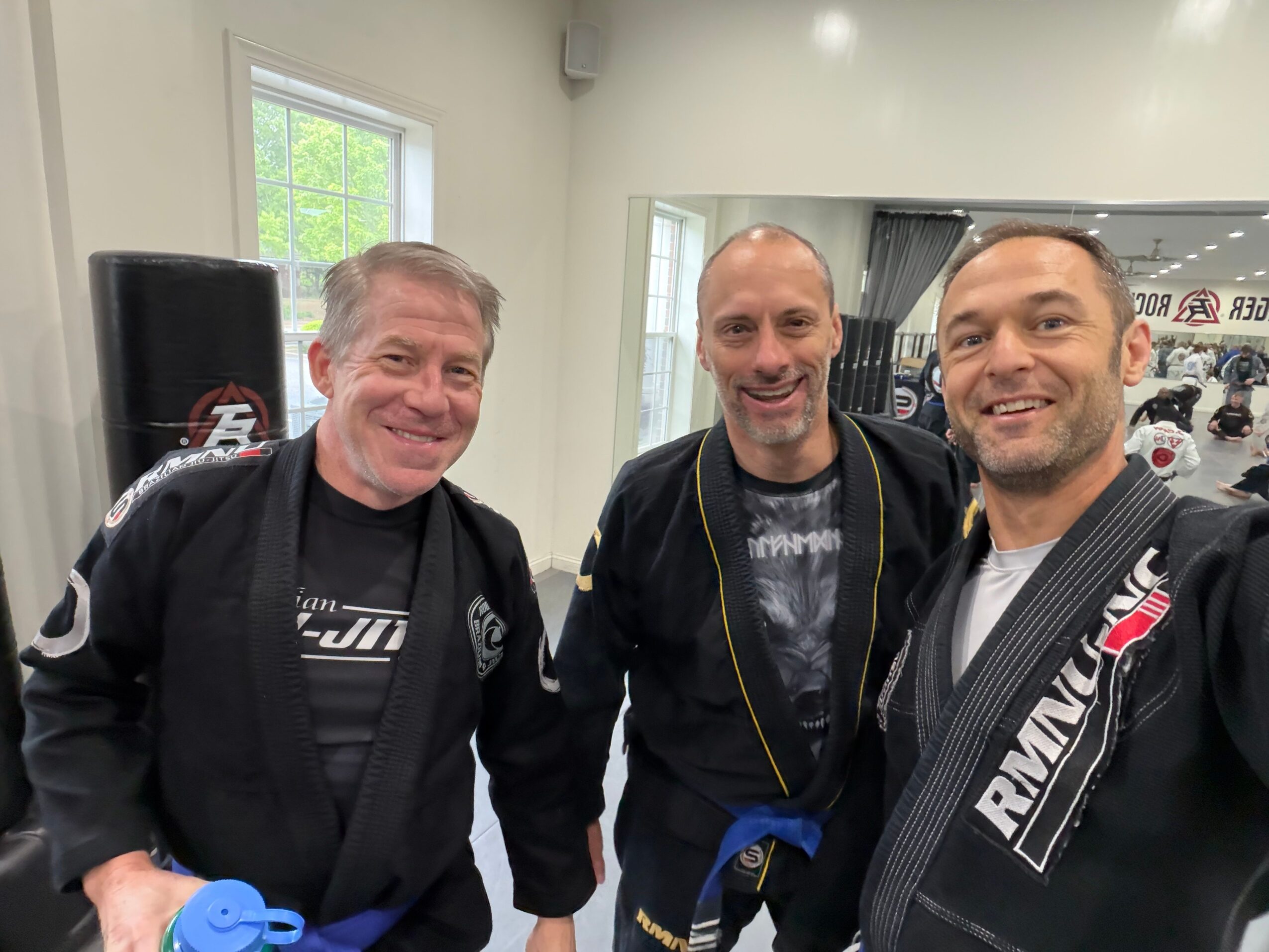 Three men posing indoors, smiling, with a window and various items in the background.