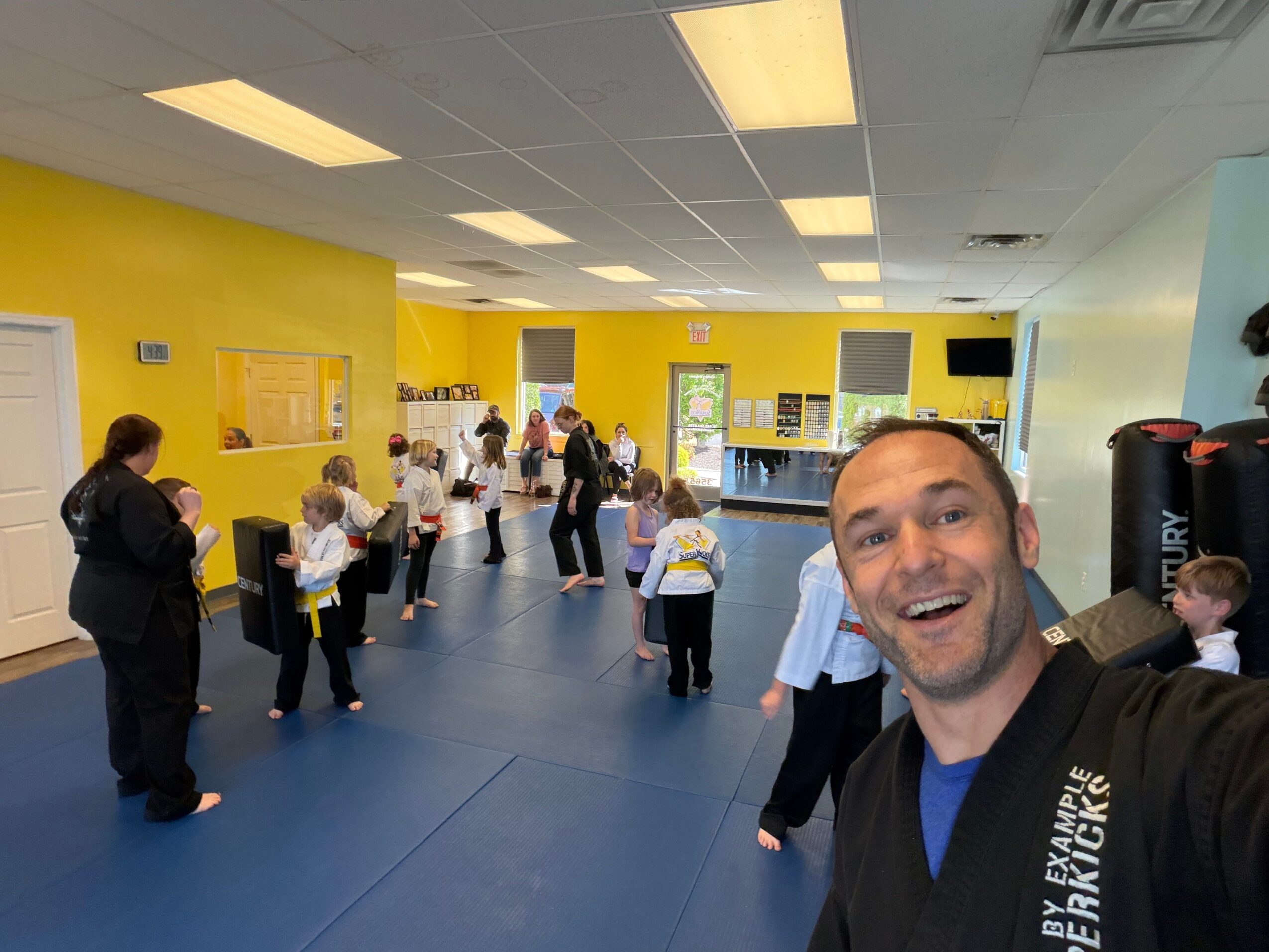 Group of children and adults in a yellow-walled room, some holding papers, with a man taking a selfie in the foreground.