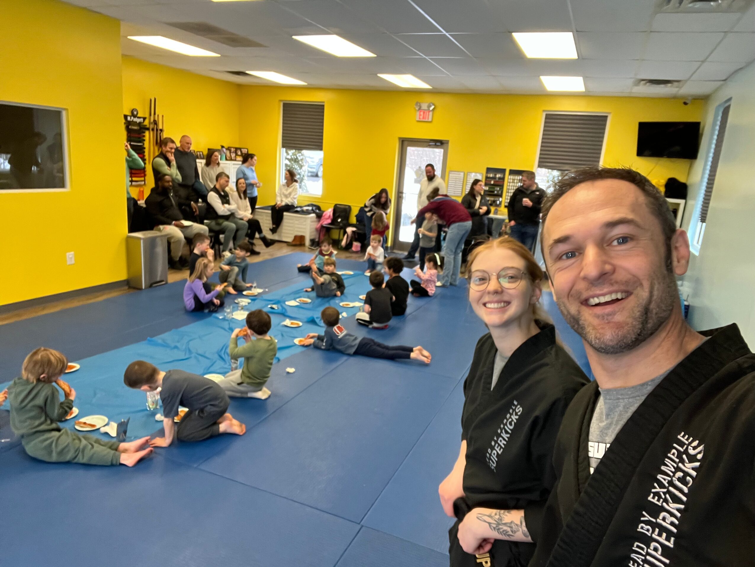 Two smiling adults taking a selfie in a brightly lit room with children sitting on blue mats, engaging in activities, yellow walls, and windows.