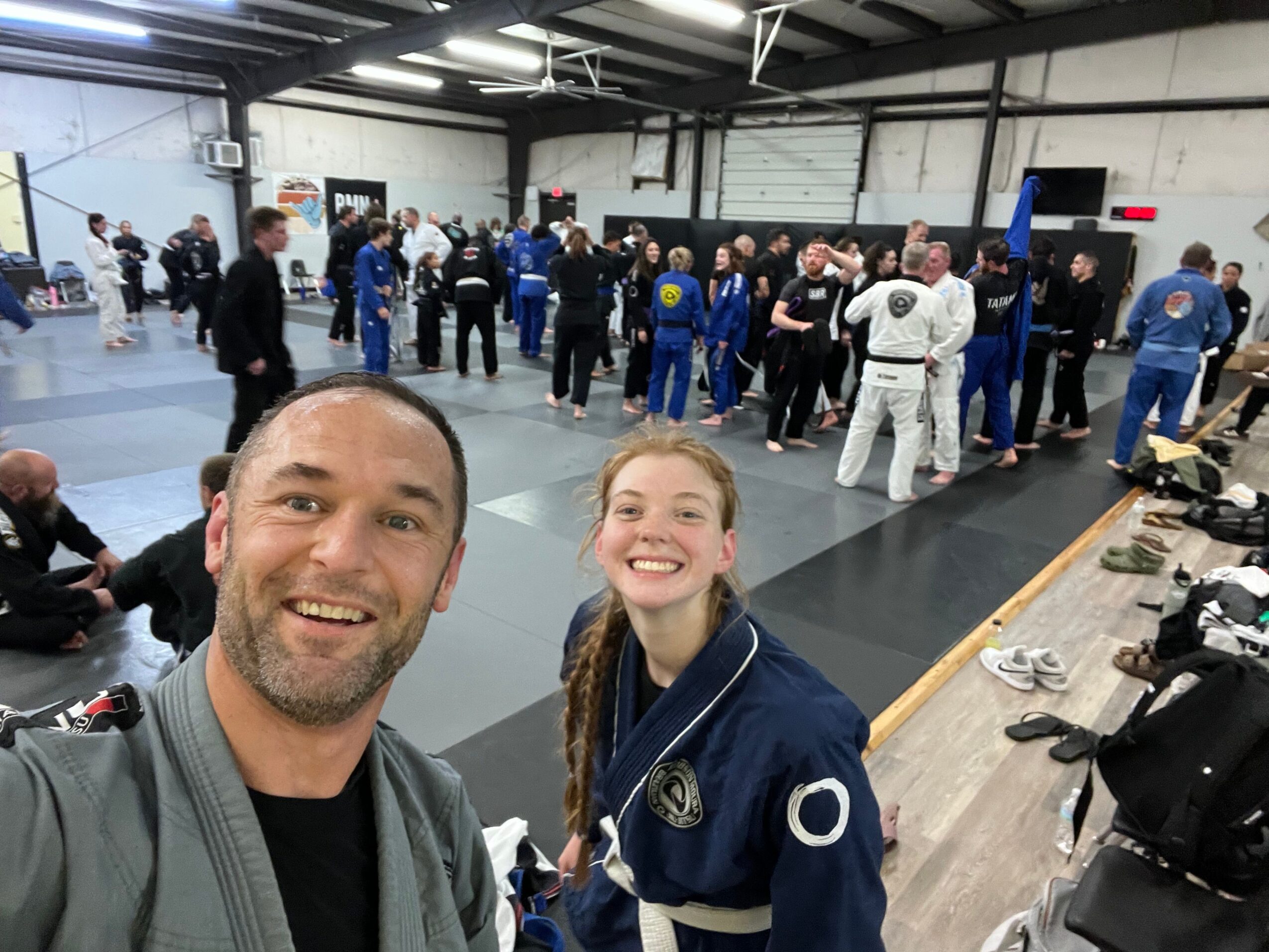 Two people taking a selfie at an indoor event with many attendees in the background.