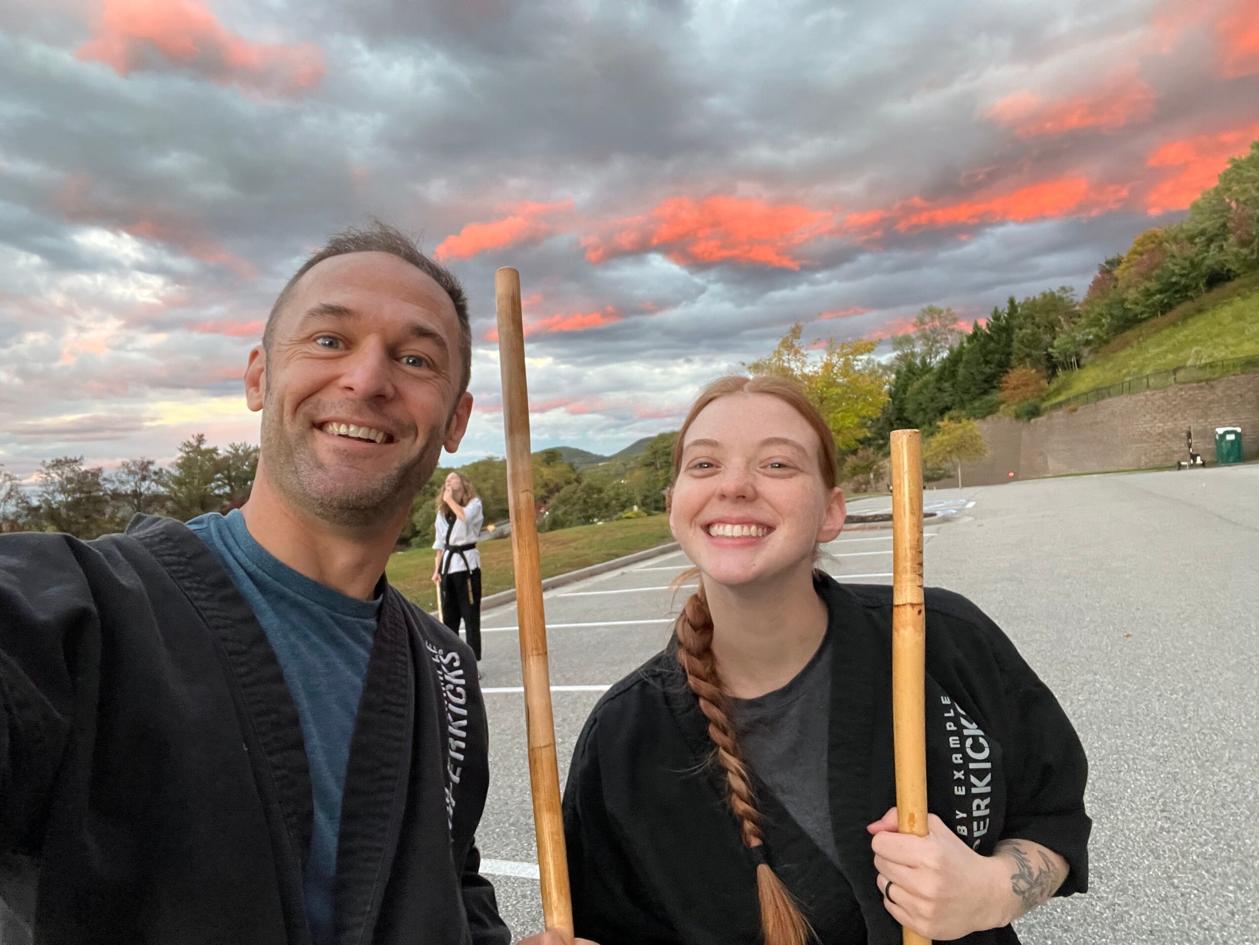 Two people outdoors holding wooden sticks, smiling, with a colorful sky and landscape in the background.