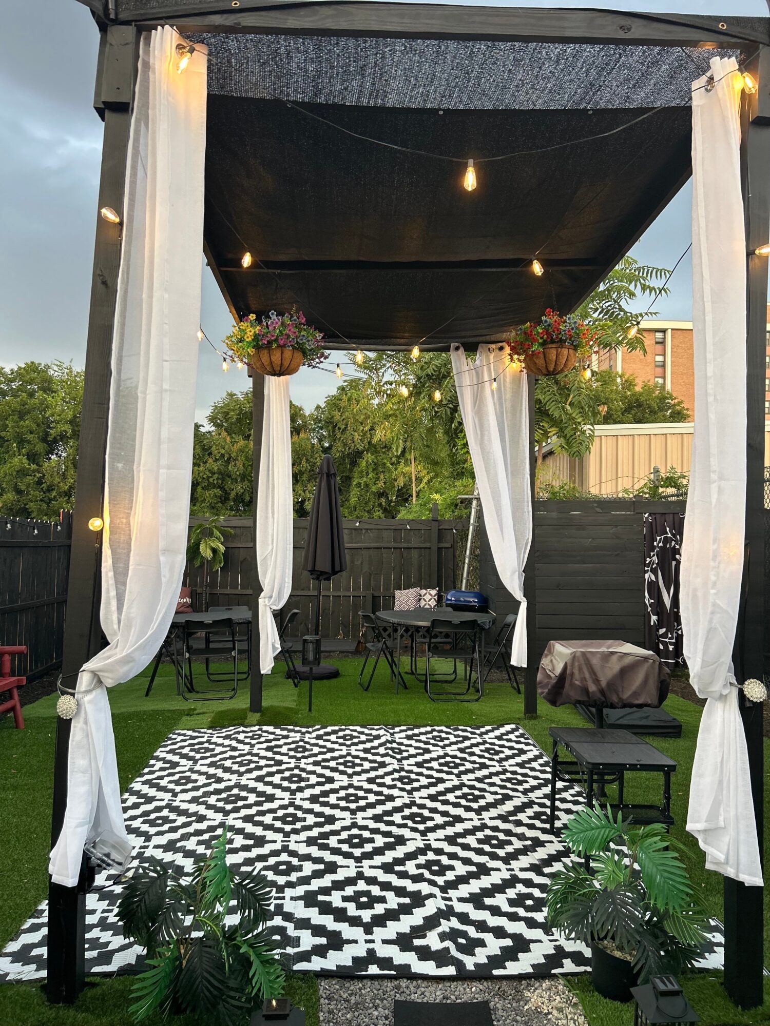 Outdoor gazebo with white curtains, string lights, and a patterned rug on grass, surrounded by trees and a fence.