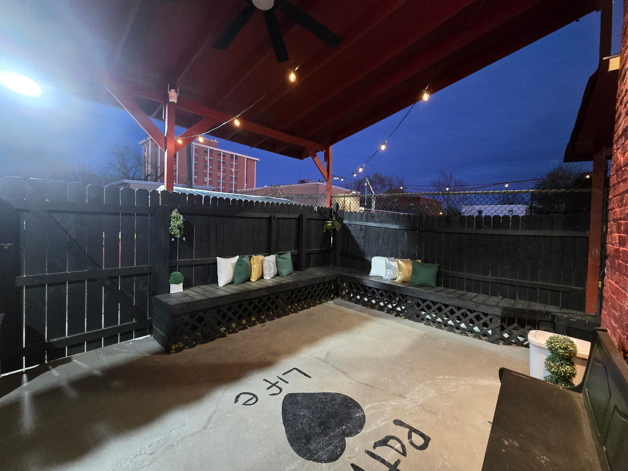 Covered outdoor patio with black fence, string lights, and a concrete floor with a heart and text, at dusk.
