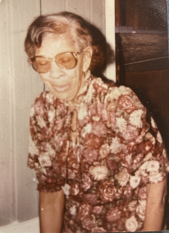 Elderly woman wearing glasses and floral dress, standing indoors, looking down, with a neutral background.