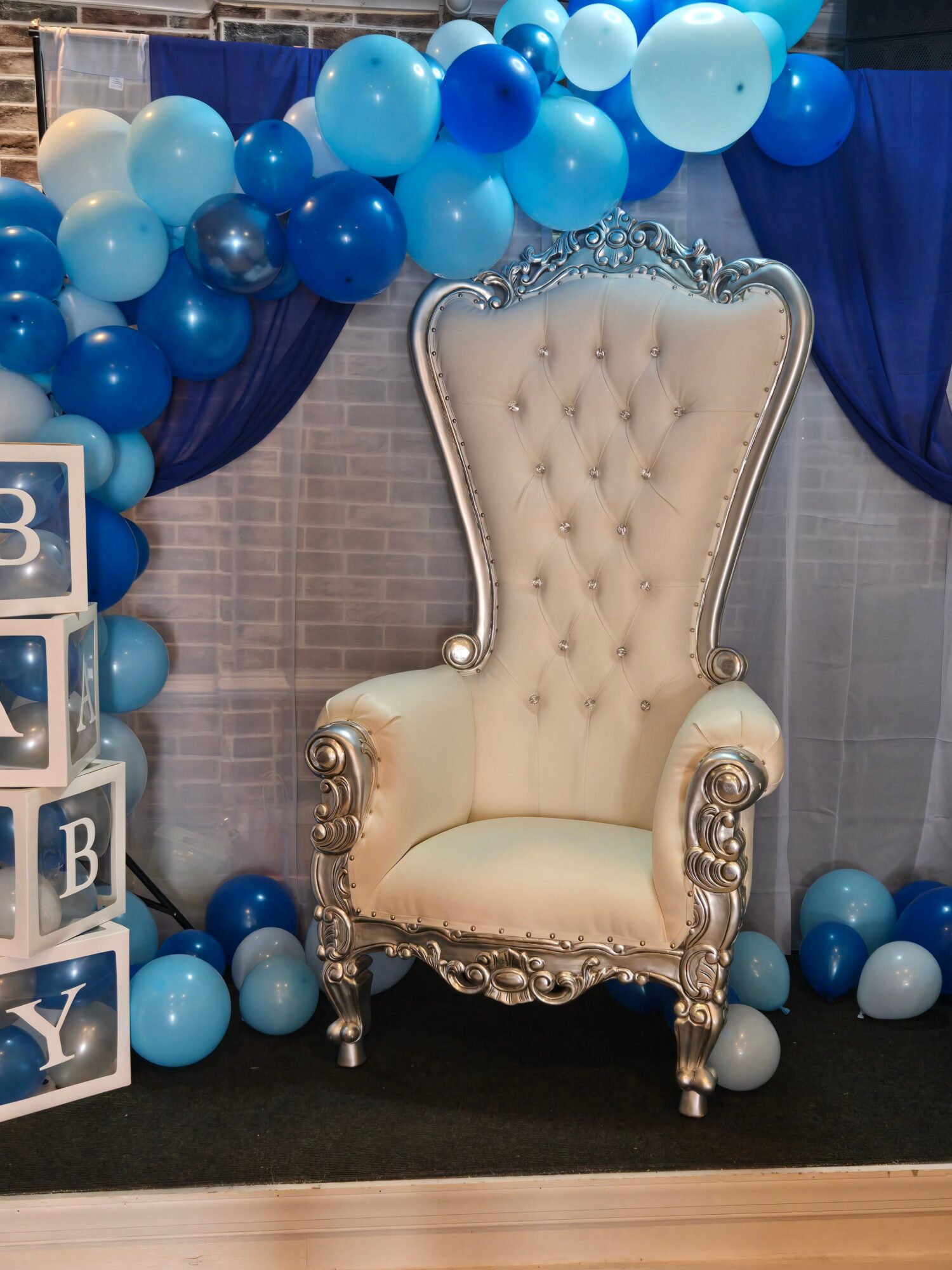 Upright ornate cream-colored throne with silver accents, surrounded by blue and white balloons, on a gray background.