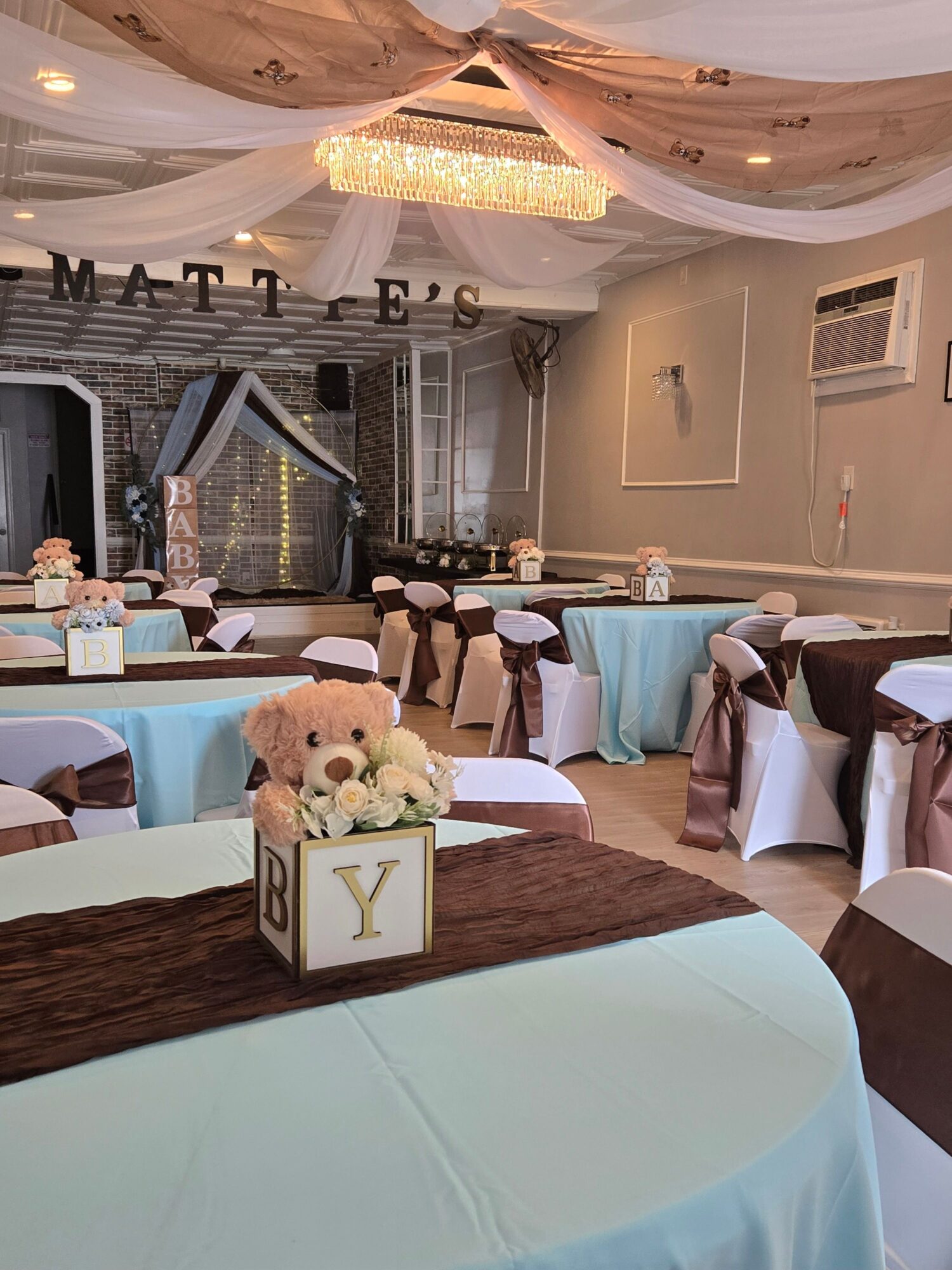 Wedding reception table with a cake, floral centerpiece, and decorated chairs in a decorated hall.