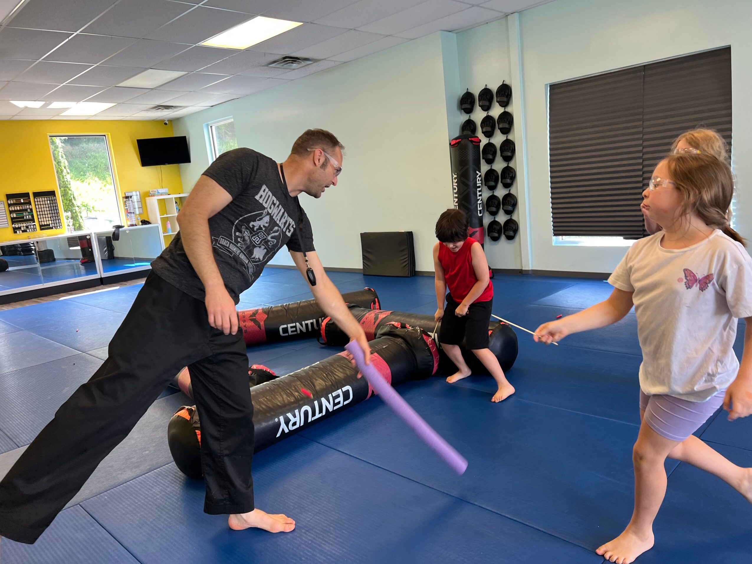 Instructor demonstrating a martial arts move with children in a training room, blue mats, and equipment in background.
