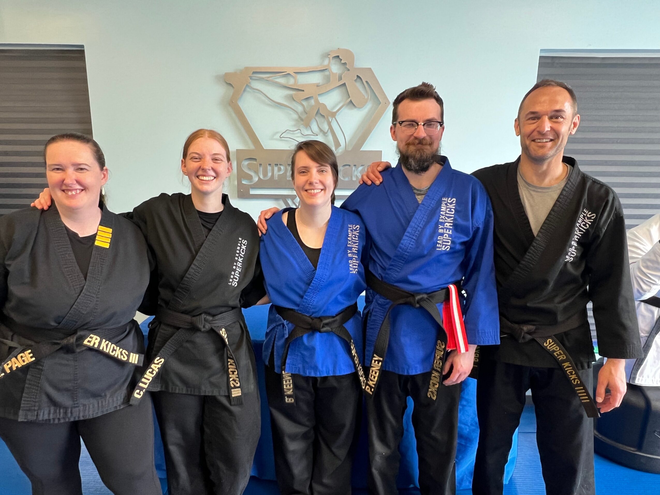 Group of five people in martial arts uniforms smiling indoors, with a sign and a skeleton model in the background.