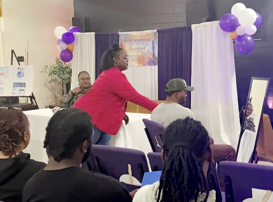 Person in red jacket speaking to audience in a decorated event space with purple and white balloons.