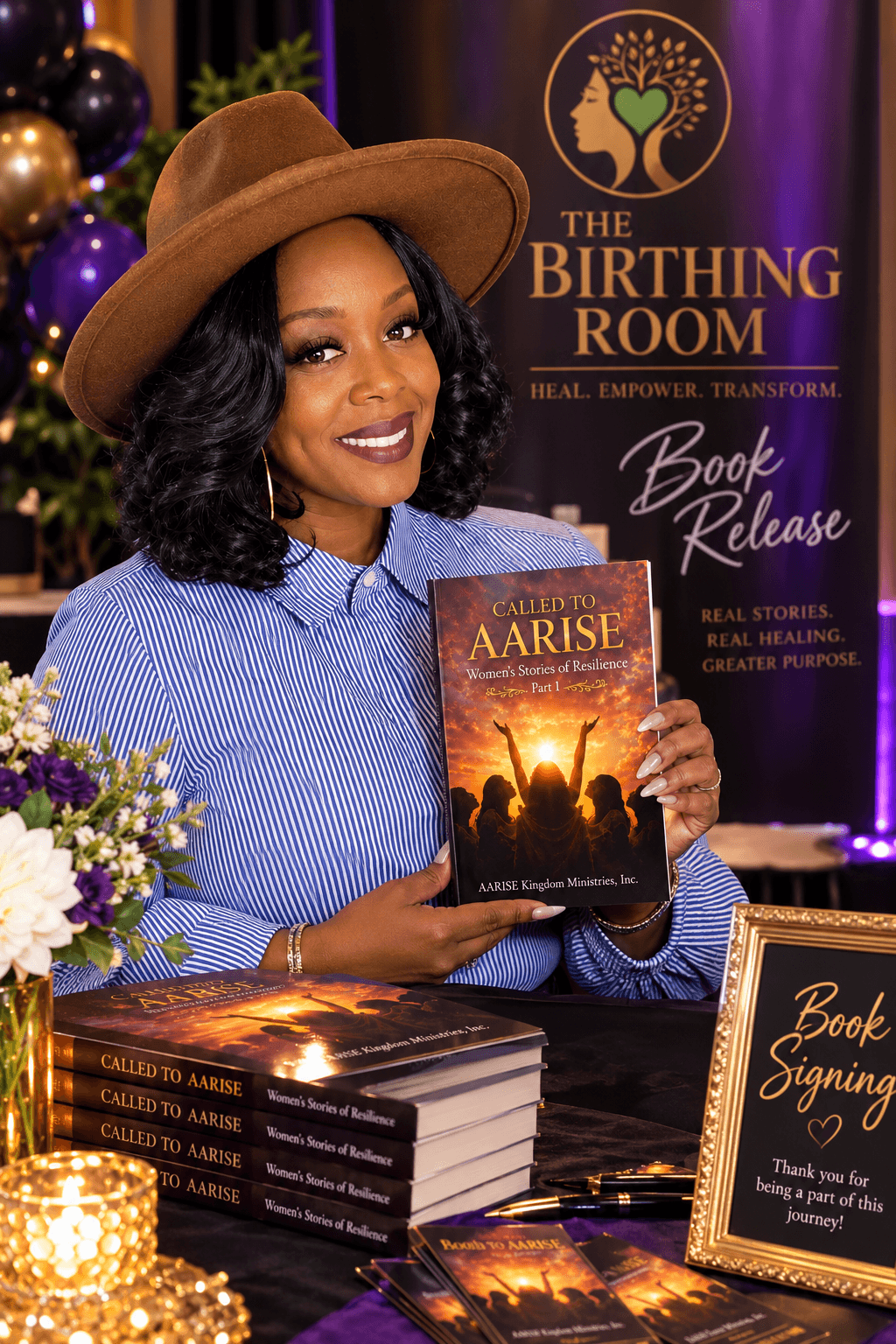 Woman with dark curly hair and a wide-brimmed hat holding a book at a book launch event, with stacked books and a floral arrangement nearby.
