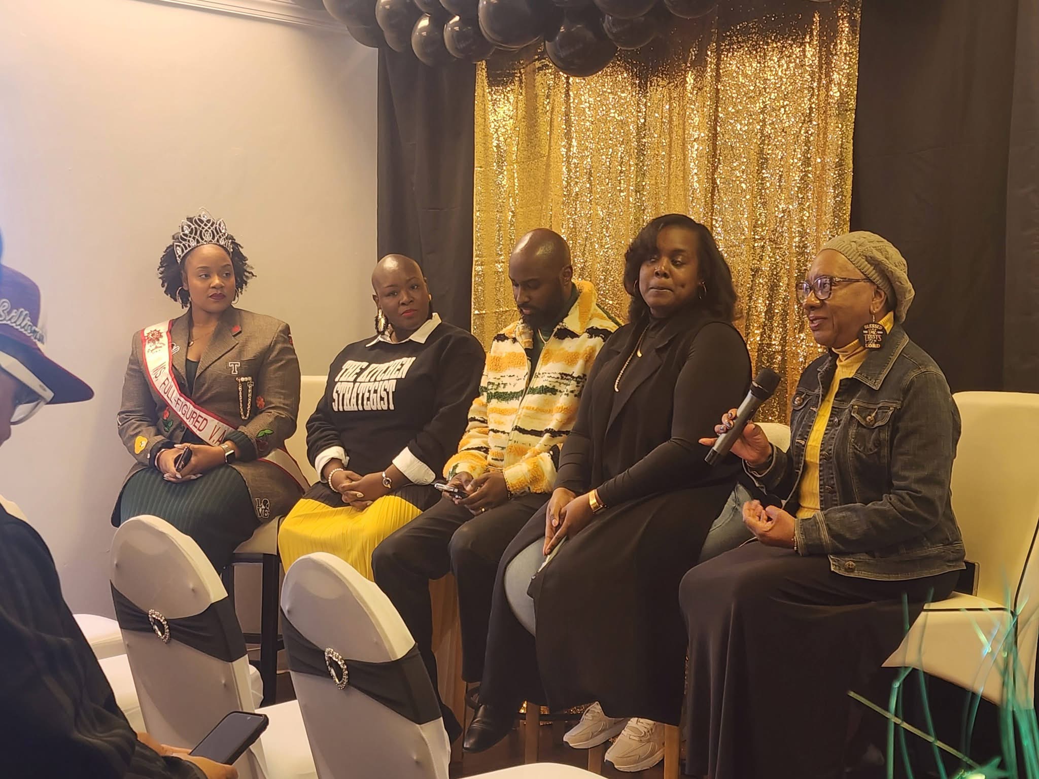 Five women seated on stage, one holding a microphone, with a gold curtain backdrop.