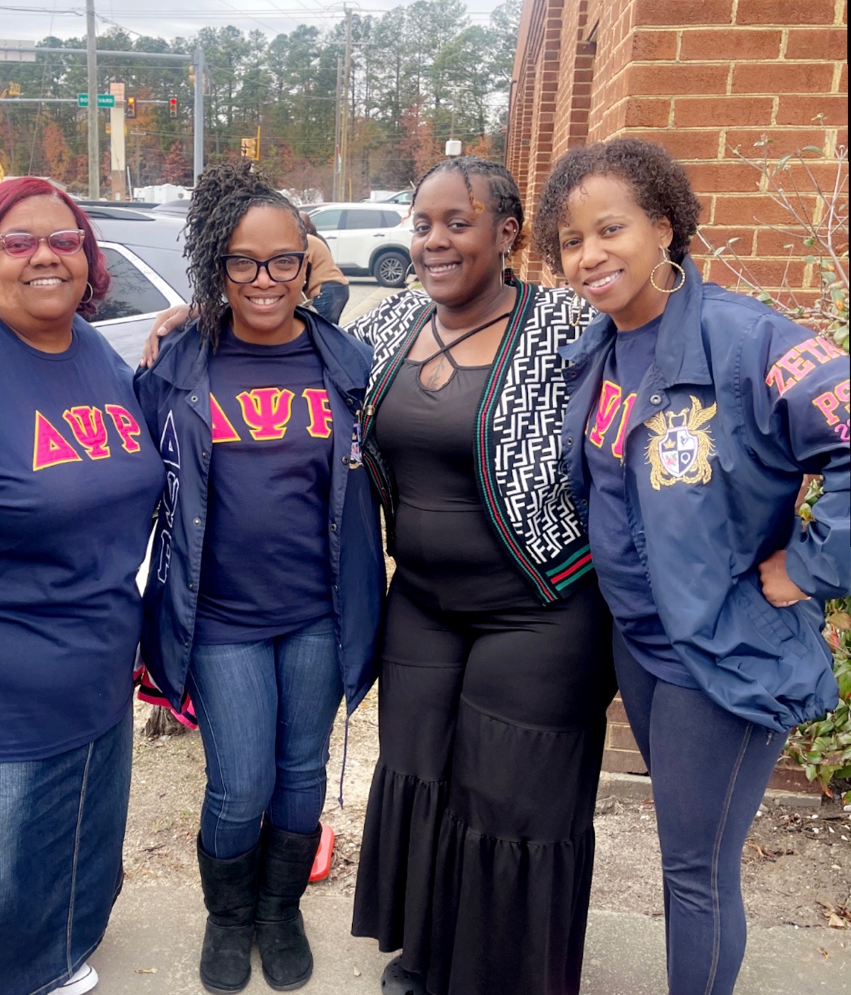 Four women standing outdoors near a brick building, smiling, wearing jackets and casual clothing.